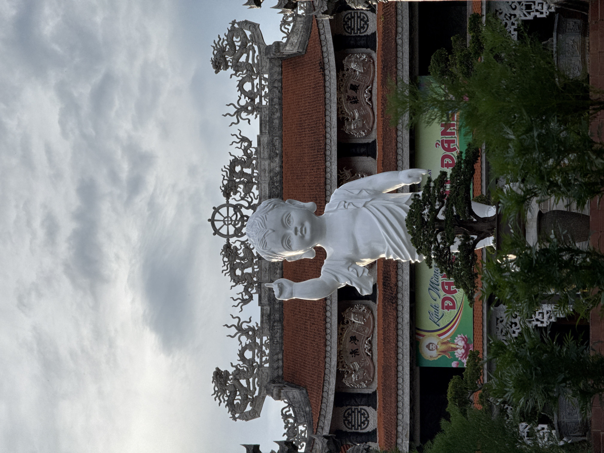 The image shows a statue of a young person, perhaps a child, made of white material, standing in front of an ornate building in Thôn 5, Vietnam. The statue is pointing with its right hand, and it is positioned in front of the building's roof, which is decorated with dragon motifs. The building has red tile roofing and ornate carvings. There is a green sign on the building to the right of the statue. The sky is overcast, and there are trees to the right of the building.