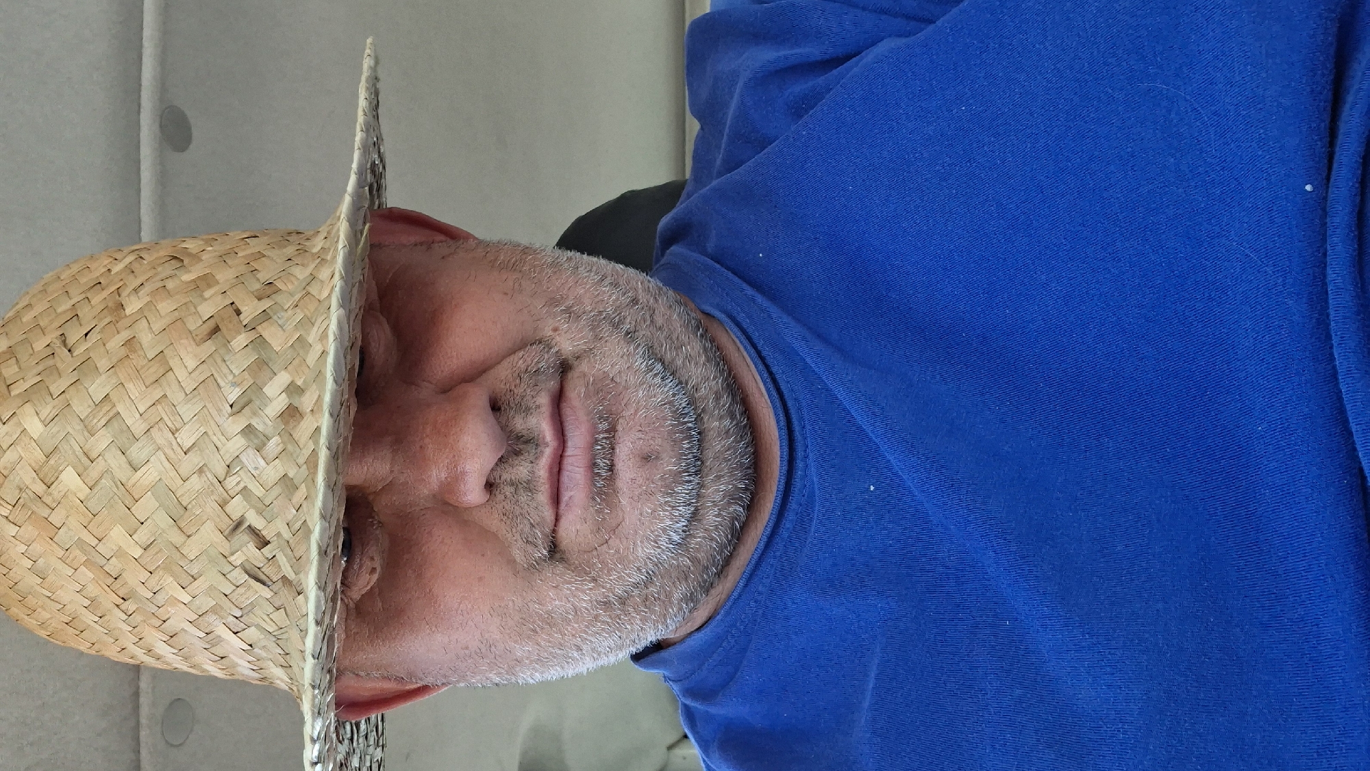 A man is wearing a straw hat and a blue t-shirt. The man is looking down at the camera. The hat is casting a shadow over his eyes, so only his nose and mouth are clearly visible. The man has a beard and a double chin. The background of the image is blurred, but it appears to be indoors. The image may have been taken in Hîncești, Moldova.