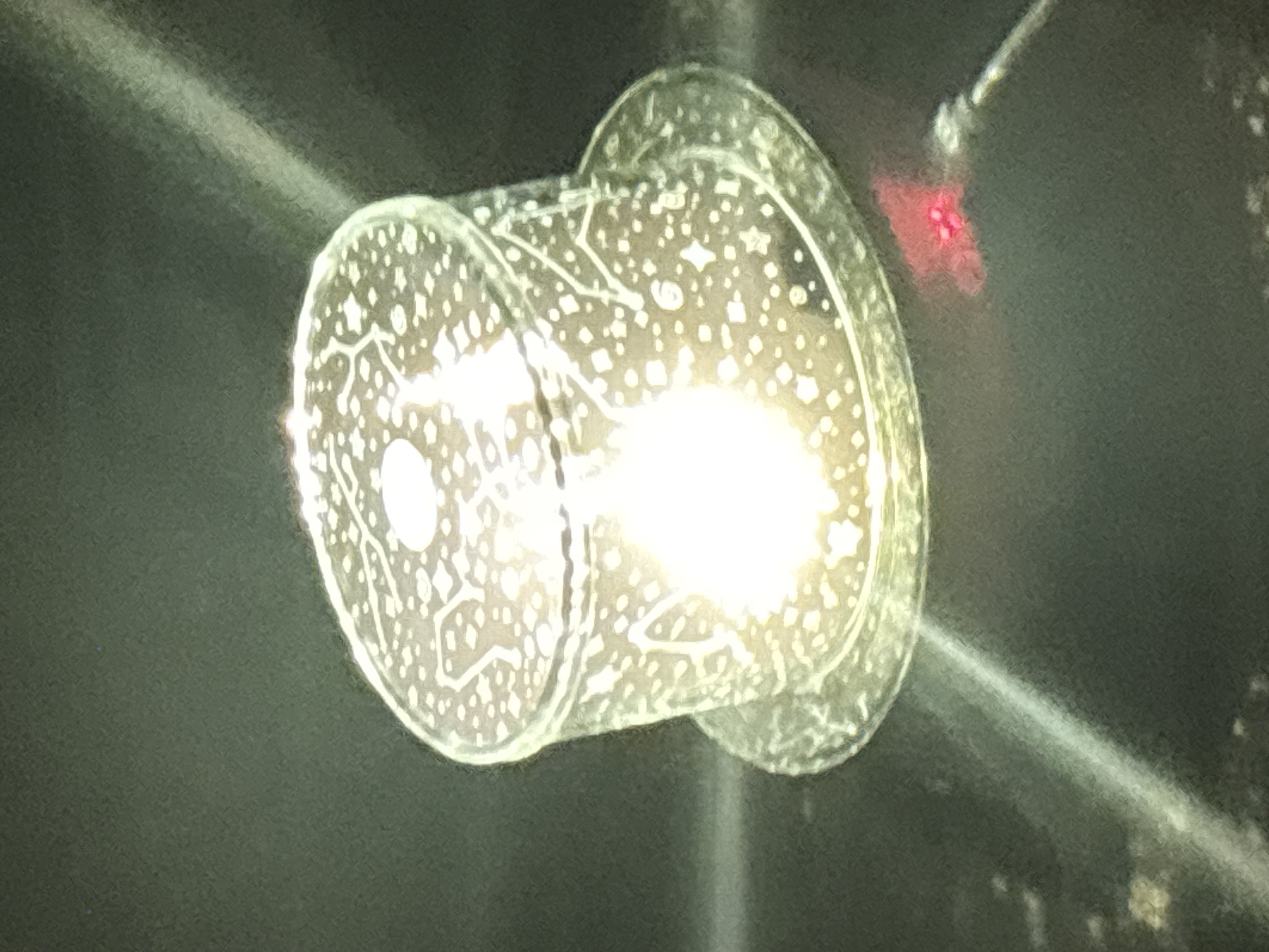 The image shows a close-up, slightly out-of-focus view of a glass object that appears to be a decorative candle holder or a small vase. It has a cylindrical shape with a flared rim and a wider base. The glass is translucent and decorated with a pattern of white dots and star-like shapes. Inside the object, a bright light source is visible, possibly a candle or an LED light, which causes the glass to glow and creates a shimmering effect. The background is dark and textured, suggesting an indoor environment. There is a faint red light source visible in the lower right portion of the image, possibly from a laser pointer or a small electronic device. The overall impression is one of ambient lighting and decorative illumination. There are no discernible elements that would place this image specifically in Kryvyi Rih, Ukraine.