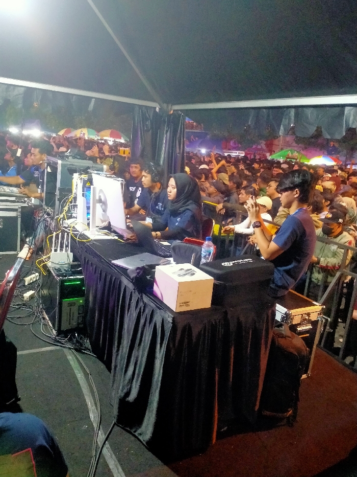 The image shows a scene taken at night under a large tent, possibly at an outdoor event. The location is in Boyolali, Indonesia, specifically at the coordinates -7.535 latitude and 110.601 longitude. A group of people are gathered around a table, working on a laptop computer and other electronic equipment. There is a large black curtain behind the table and numerous cables are seen running across the floor. Three individuals are clearly visible: a man in a blue shirt, a woman wearing a hijab, and another man seated in a chair facing away from the camera. The scene appears to be a technical area, possibly related to live audio or video production, at an event in Boyolali.