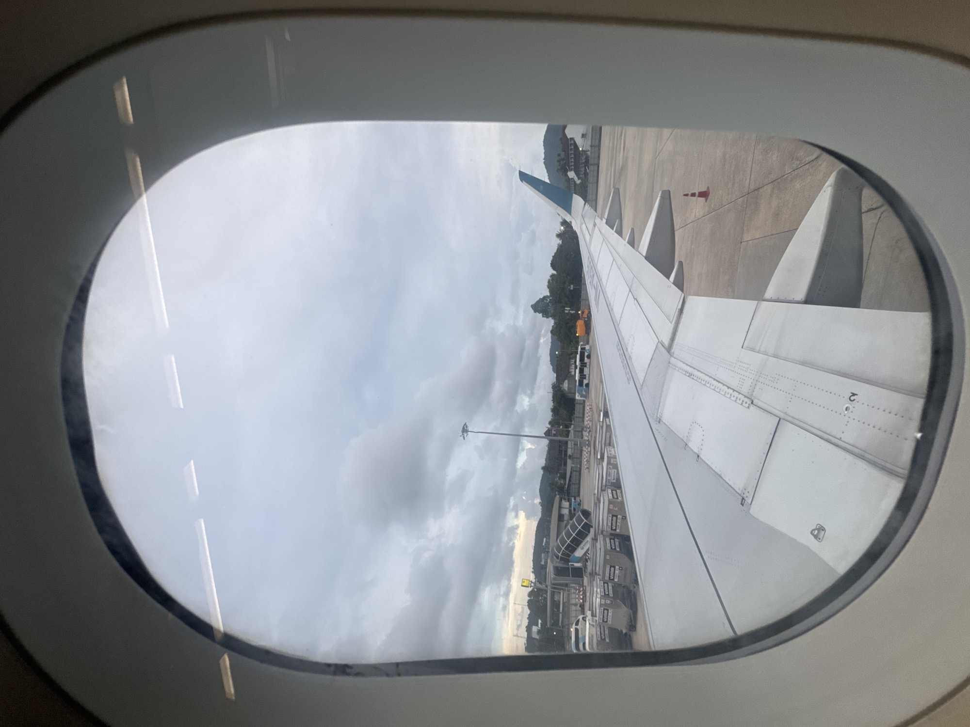 The image, taken from an airplane window, shows an airport tarmac at Tambon Mai Khao, Thailand. The view encompasses a portion of the aircraft's wing in the foreground. Beyond the wing, the tarmac is visible with various objects such as cargo containers, a jet bridge, service vehicles, and a traffic cone. In the background, there are buildings and lush green hills under a cloudy sky. The lighting suggests it is either early morning or late afternoon. There are no visible people in the frame.