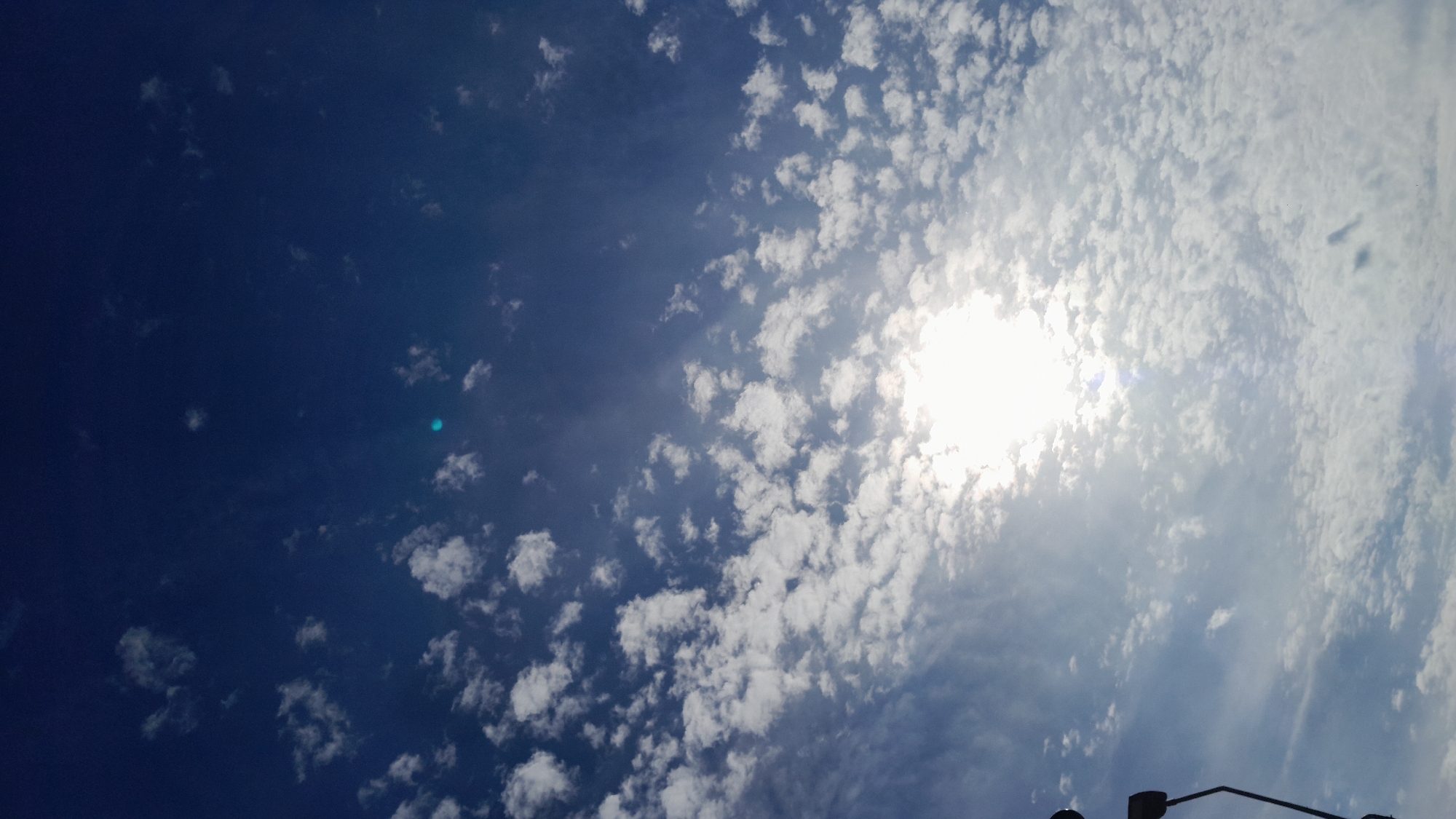 A vertical, low-angle shot captures a bright, sunny sky filled with a scattered pattern of white, puffy clouds. The sun is a dominant element in the frame, appearing as a brilliant white orb positioned in the lower-middle section of the image, emitting a strong glare that washes out details in its immediate vicinity. The sky itself is a deep, rich blue, providing a stark contrast to the white clouds and the intense sunlight. The scene is reminiscent of a clear day, with the sun's rays filtering through the thinner parts of the cloud cover, creating a bright and airy atmosphere. In the bottom left corner, a dark, silhouetted object is visible, appearing to be the arm of a street lamp or a similar fixture, extending upwards into the sky. The overall impression is one of a serene and expansive sky, characteristic of good weather in an outdoor setting, potentially a park or open area within Krakow, Poland, given the implied urban context.