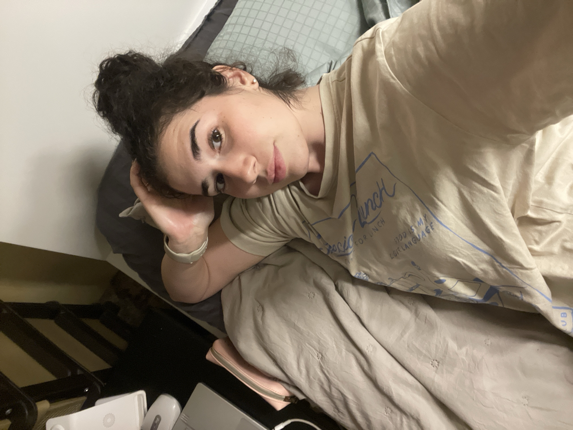 A woman with dark curly hair partially gathered in a bun is lying on a bed. She wears a beige t-shirt with a blue design, her head resting on her hand. The bed has a gray pillow and a beige patterned blanket. In the lower left corner, there is a dark wooden structure with various white electronic devices. It's not possible to determine if this photo was taken in Sofia, Bulgaria or determine a location.