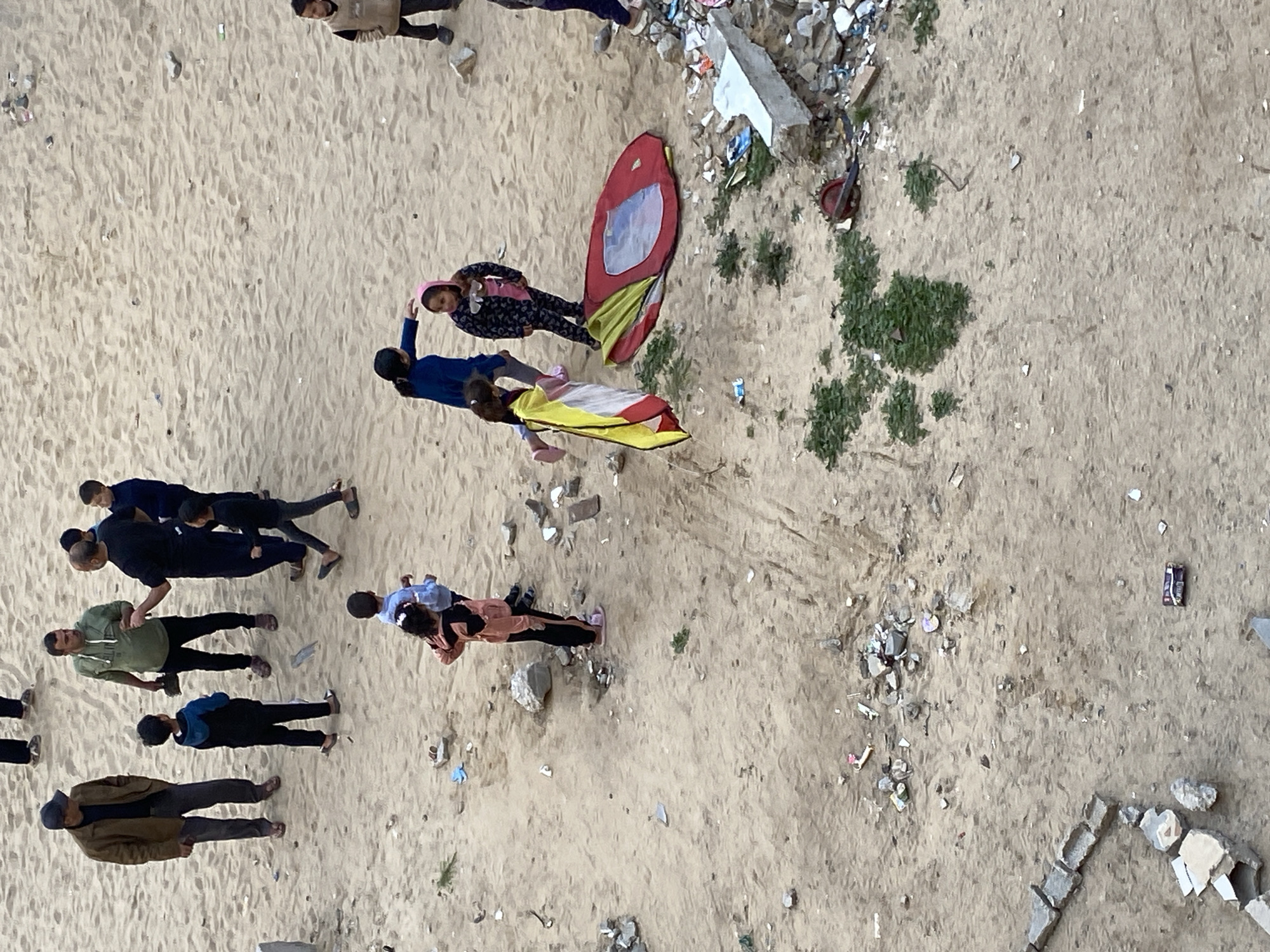 The scene shows a group of people standing on a sandy area, possibly a beach or a vacant lot in Khan Yunis, Palestinian Territory. Debris and scattered trash are visible on the ground. The group is composed of adults and children. Two children are interacting with what appears to be small pop-up tents, one red and one yellow. The adults are standing nearby, some in groups, others alone. Some are dressed in casual attire, including pants and t-shirts. The area shows evidence of urban decay, with exposed soil, scattered rubble, and refuse. The image appears to have been taken from an elevated perspective, looking down on the group.