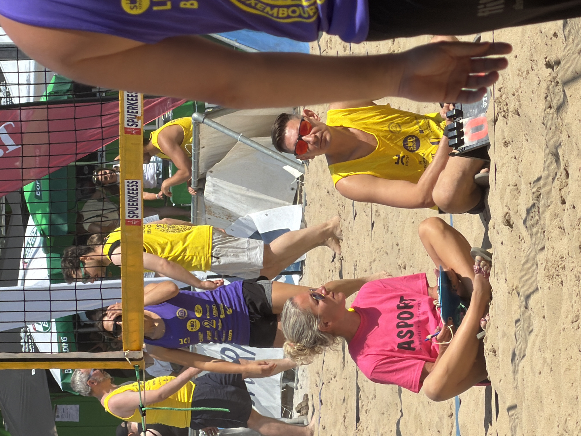 The image depicts an outdoor beach volleyball event, likely taking place in Esch-sur-Alzette, Luxembourg, as indicated by the presence of "SPUERKEES" banners, a known sponsor associated with the country. The scene is set on a sandy court, with a net dividing the playing area.
In the foreground, a woman wearing a bright pink t-shirt is seated cross-legged on the sand, holding a clipboard and pen. To her right, a man in a yellow tank top and red sunglasses is also seated, looking towards his left.
In the middle ground, behind the net, a woman in a purple tank top and black shorts is standing, her hands near her face. Next to her, a man in a yellow tank top and light-colored shorts is also standing. Further to the left, another man in a yellow tank top and dark shorts is standing.
The background shows various structures, including tents, umbrellas, and more people, suggesting a public event. The overall atmosphere appears to be that of a sporting competition on a sunny day.
