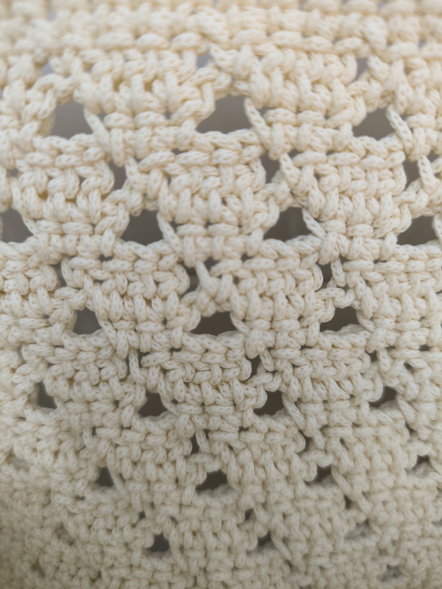 This is a close-up, vertical shot of cream-colored crochet fabric. The pattern consists of repeating diamond shapes with open spaces in between. The texture is thick and slightly fuzzy, characteristic of yarn. The lighting is even and diffused, highlighting the intricate stitch work. The image is cropped tightly on the fabric, with no background elements visible, making it impossible to determine a location in Krasnaya Polyana, Russia or any other context. There are no people, actions, or interactions depicted in the image.