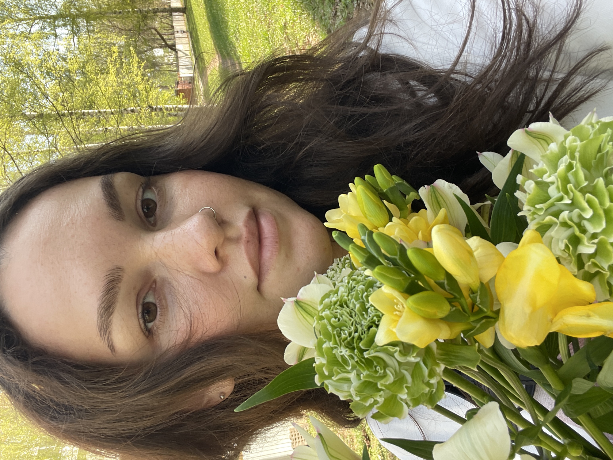 A woman with long brown hair and a nose ring is holding a bouquet of yellow and green flowers. The background shows trees and a grassy area. The image does not contain information to identify the location as the city of Izhevsk, Russia.