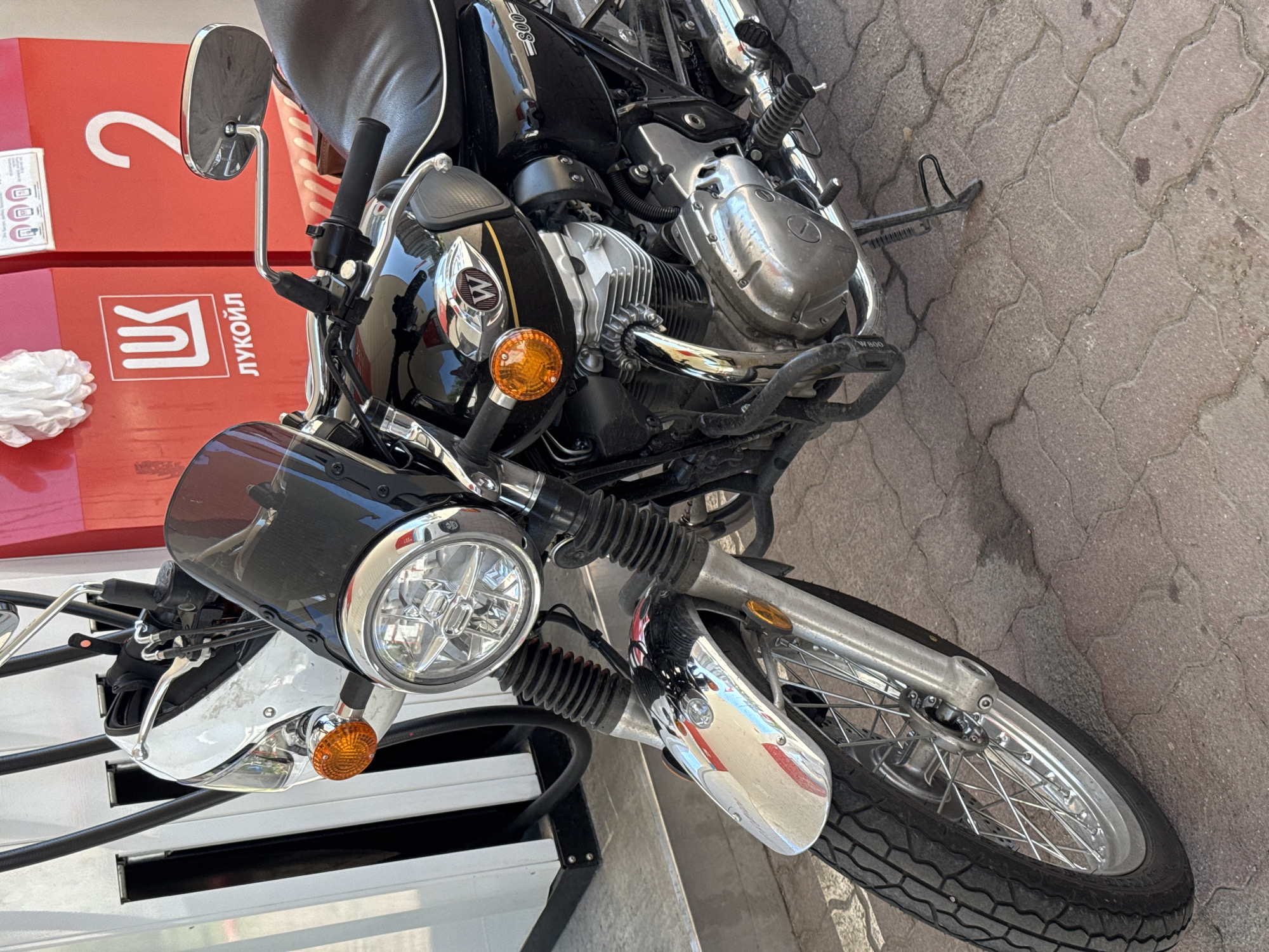 A classic-style motorcycle is parked at a Lukoil gas station in Sinyavino, Russia, captured from an elevated, angled perspective. The motorcycle, predominantly black, features numerous chrome details including the engine block, headlight bezel, front fender, mirrors, and spoked wheels. The gas tank bears a stylized 'W' logo and the text "W 800". An orange turn signal is visible near the headlight.
In the background, a bright red gas pump or dispenser prominently displays the Cyrillic logo "ЛУКОЙЛ" (Lukoil) and a large white number "2." A crumpled white object, possibly a paper towel or glove, rests on the pump. The ground is paved with reddish-brown interlocking stone bricks. The bright illumination suggests it is daytime. No people are visible, but the motorcycle appears to be stationary, indicative of a fueling stop.