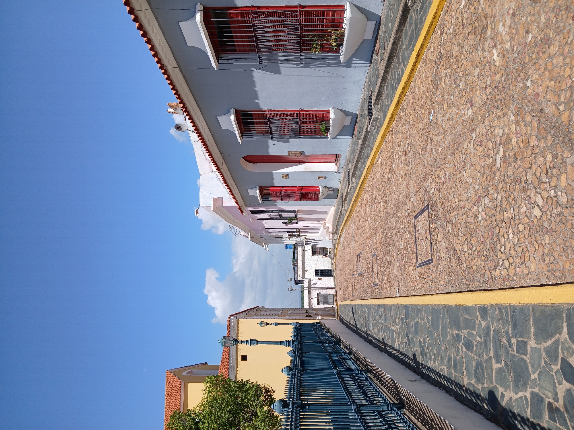 The image depicts a street scene in Ciudad Bolivar, Venezuela, characterized by colonial architecture. Buildings lining the street are painted in various colors, including blue, white, pink, and yellow. Many of the buildings have red window and door frames, some with decorative metal grilles and window boxes. The street is paved with cobblestones, divided by a yellow stripe marking the sidewalk. A blue metal fence borders the lower portion of the image, with street lamps positioned along it. The sky is clear and blue with a few scattered clouds. A tree partially obscures the lower left corner. The scene appears to be taken during the day.