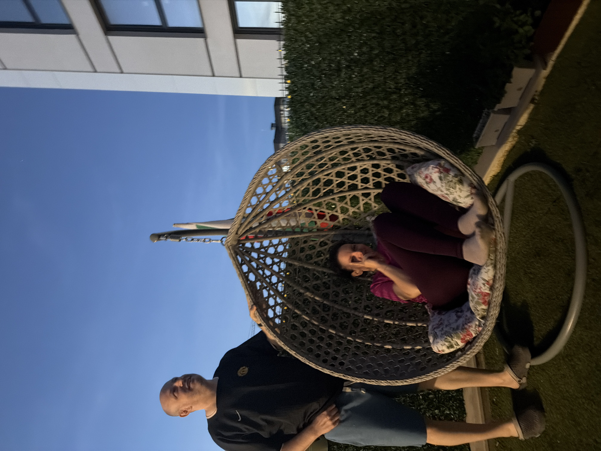 A man stands to the left of a round, woven hanging chair in which a woman is seated. The man is bald, wearing a black t-shirt and blue shorts. The woman is wearing a purple shirt and dark pants. She is looking at her phone and holding up two fingers. The hanging chair has floral cushions. The background consists of a green hedge, a modern building, and a clear blue sky, indicating it is dusk. The ground appears to be artificial grass.