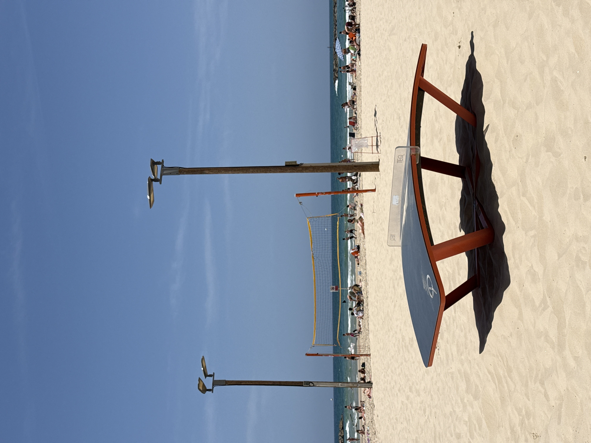 The image depicts a beach scene in Tel Aviv, Israel, under a clear blue sky with scattered clouds. The foreground is dominated by a beach volleyball net and a TEQ table tennis table, casting a shadow on the sand. Wooden lampposts line the beach. In the background, people are scattered along the shoreline, some swimming in the water. The beach sand is light in color.