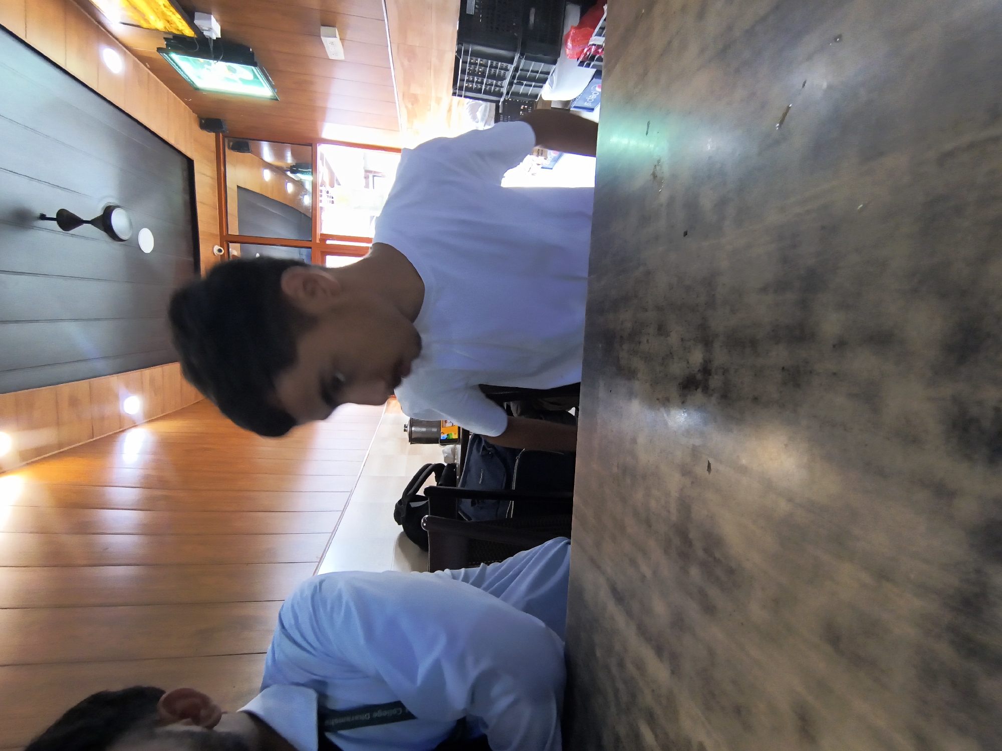 The image depicts two young males in what appears to be a casual indoor setting, possibly a restaurant or cafe in Dharamshala, India. The foreground is dominated by a wooden table with a textured surface, reflecting some light. In the midground, the person on the right, wearing a white t-shirt, is looking downwards and to the left, with their face partially obscured. To the left, another person, dressed in a light blue shirt with a lanyard bearing the text "College Dharamsala", is visible from the chest up, looking towards the left.
The background reveals a wall paneling made of wood, with lighting fixtures embedded in the ceiling. A ceiling fan is visible overhead. Further back, there is a doorway or passage, possibly leading to another section of the establishment, with natural light streaming in. Several black plastic crates are stacked against the back wall, along with some white containers and a red item, likely packaging. A television screen displaying some green content is mounted on the wall. The overall impression is a candid shot within a public space in Dharamshala.