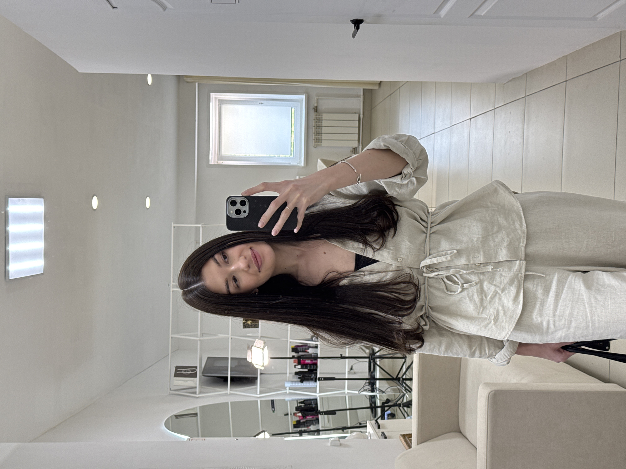 A woman takes a selfie in a bright, white room. She wears a light beige linen-like outfit. Her dark hair is long and straight. She holds a black smartphone up to take the picture. Behind her is a salon or studio space with lighting equipment, shelves, and a large circular mirror on the wall. A window is visible in the background. The location may be Chaikovsky, Russia, though this is not visually confirmed.