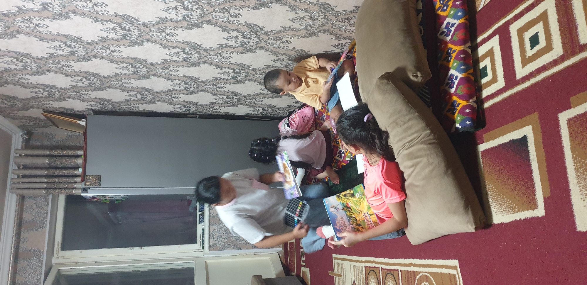 The image depicts a domestic scene indoors, likely a living room in Fergana City, Uzbekistan. Four individuals are present: an adult in a white t-shirt and blue jeans is kneeling and holding a book and a marker set. To their right, a young girl with dark hair in pigtails is also kneeling, facing away from the viewer, and appears to be looking at a book. Further to the right, a young boy in a yellow polo shirt and blue shorts is sitting and looking down at a book. In the foreground, a young girl in a pink t-shirt is lying on her side on cushions and looking at a book. The room has patterned wallpaper and a red carpet with geometric designs. A large, gray refrigerator is visible in the background, along with a window and curtains. The overall setting suggests a casual, family-oriented activity, possibly a study session or playtime.