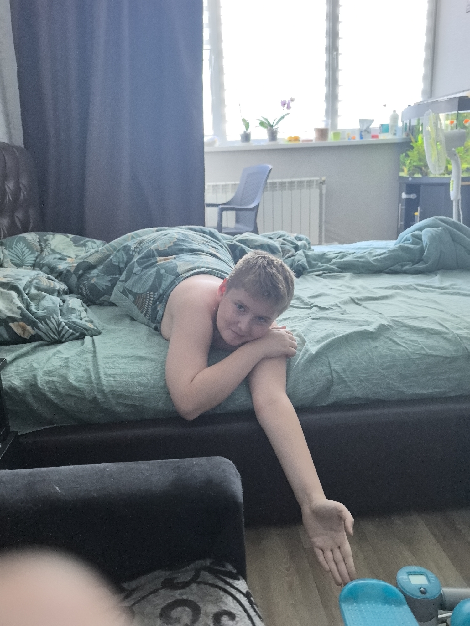 A young boy with short blond hair is lying on a bed with a teal comforter that has a leaf pattern. He is facing the camera, propped up on his elbows and looking at the viewer with a slight smile. His arms are crossed in front of him. The bed is covered with a teal sheet and a patterned teal comforter. Behind the bed, a window is visible with natural light streaming in, indicating daytime. On the windowsill, there are potted plants and various bottles and objects. To the right of the window, an aquarium with green plants and fish is visible. A white electric fan is also present. In the background, a radiator and a dark blue chair can be seen. In the foreground, a dark upholstered couch or chair is visible. The setting appears to be a bedroom. The overall mood is relaxed and casual.
