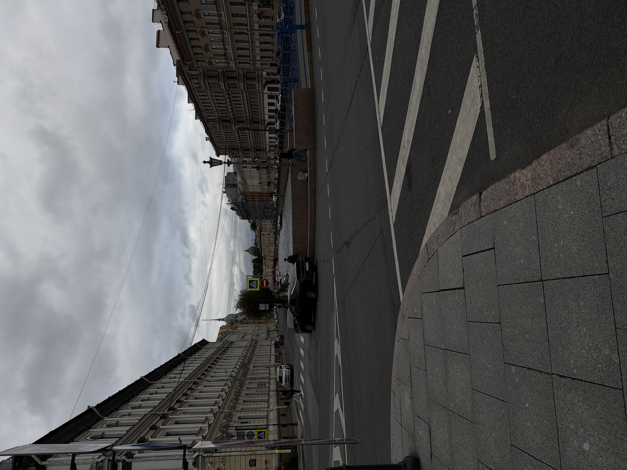 The image shows a street scene in Saint Petersburg, Russia, likely near the Neva River. A street intersection is visible with a black car driving away from the camera and a white SUV crossing the intersection. Pedestrians are present on the sidewalks and crossing the street. Buildings line both sides of the street, featuring classical architecture with multiple stories and windows. The sky is overcast with dark clouds. A blue bridge railing is visible in the background.