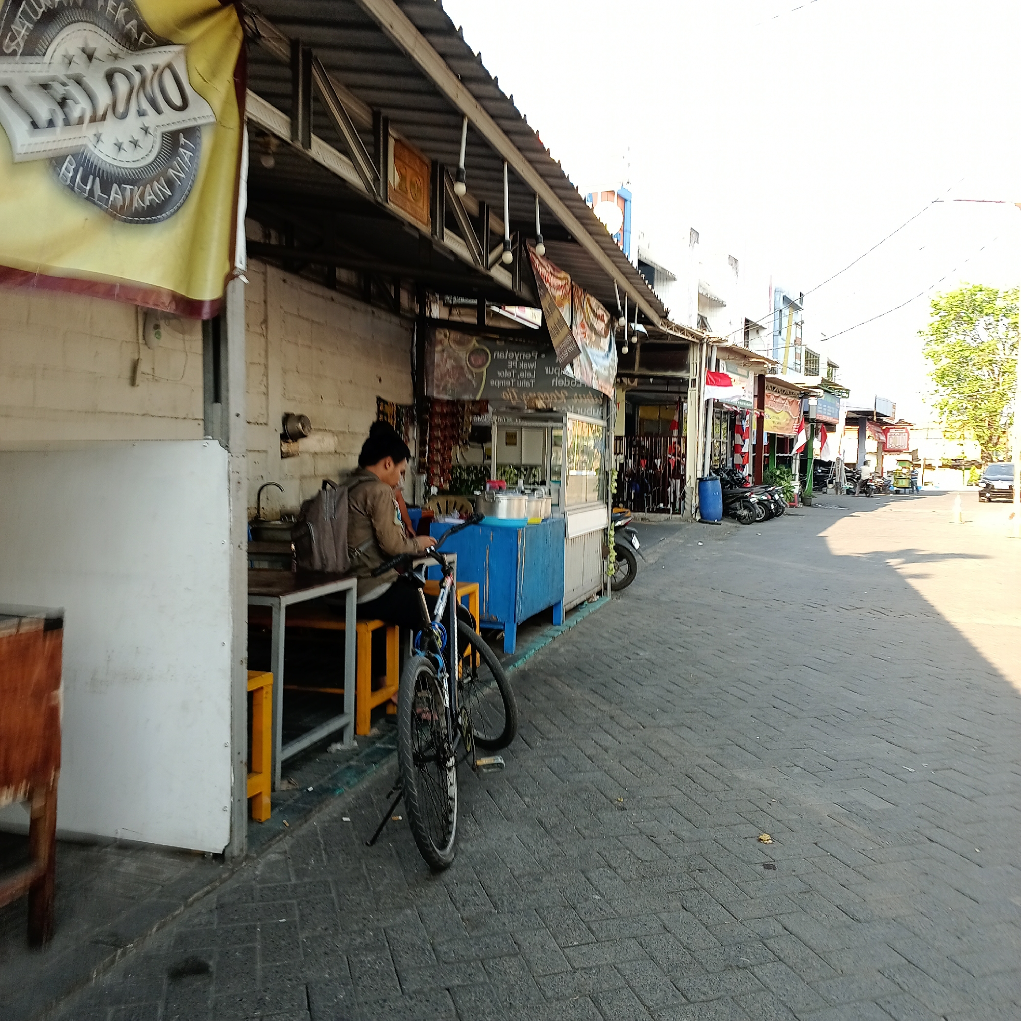 The image shows a street scene in Wedoro, Indonesia, with a focus on a small food stall. A person wearing a backpack is seated on a stool at the stall, looking down at something in their hands. A bicycle is parked next to them. The stall itself has a blue cabinet and a metal counter, with pots and other cooking utensils visible. Several other stalls and buildings line the street, with motorcycles parked in front of them. The street is paved with interlocking bricks, and the sky is bright and clear. There are flags and banners displayed on some of the stalls, indicating commercial activity. The overall impression is one of a bustling local market or street food area.