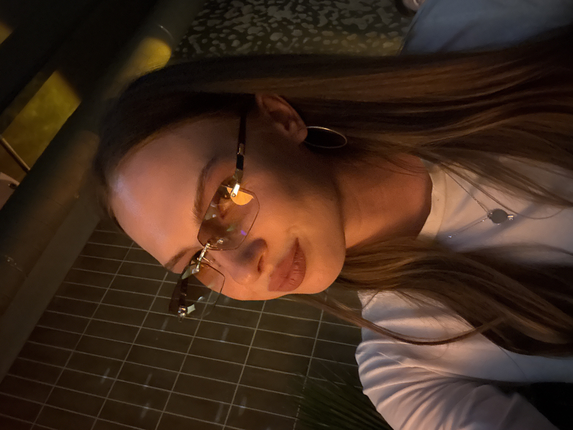 The image depicts a close-up, side-profile shot of a young woman wearing rimless sunglasses and a white t-shirt. She has long, brown hair and is wearing a silver necklace with a circular pendant and hoop earrings. The lighting suggests an indoor setting, possibly a bar or restaurant, with warm, ambient light reflecting off her sunglasses. In the background, a textured wall and a grid-like structure are visible. There is no discernible location in the city of Perm, Russia indicated in the image.