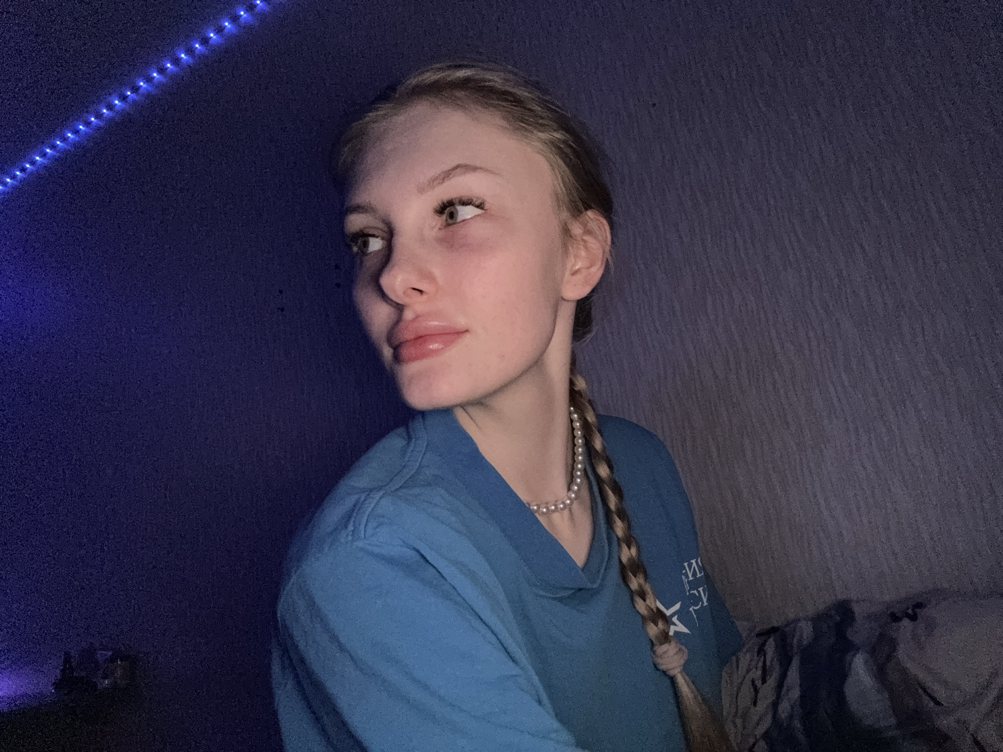 The image shows a young woman with light-colored hair pulled back and styled in a long braid, which rests over her right shoulder. She is seen in profile, looking towards her left (viewer's right) with a neutral, contemplative expression. Her features include full lips and long eyelashes. She wears a light blue or teal long-sleeved t-shirt, and a pearl necklace is visible around her neck. On the right side of her shirt, partially visible and reversed, there appears to be a design including the letter "M" and a star, suggesting a logo or text.
The setting is indoors, likely a room or confined space, dimly lit by a strong blue light source. A strip of blue LED lights runs diagonally across the upper left corner of the frame, providing the primary illumination and casting a cool, dark blue hue over the scene. The background consists of a dark, textured wall, possibly fabric or wallpaper, which appears deep blue or purple due to the lighting. A dark blanket or fabric is visible on the far right side of the image, suggesting she might be seated on a bed or soft surface. The overall atmosphere is quiet and subdued, indicative of nighttime or a low-light environment in Кондратово, Russia.