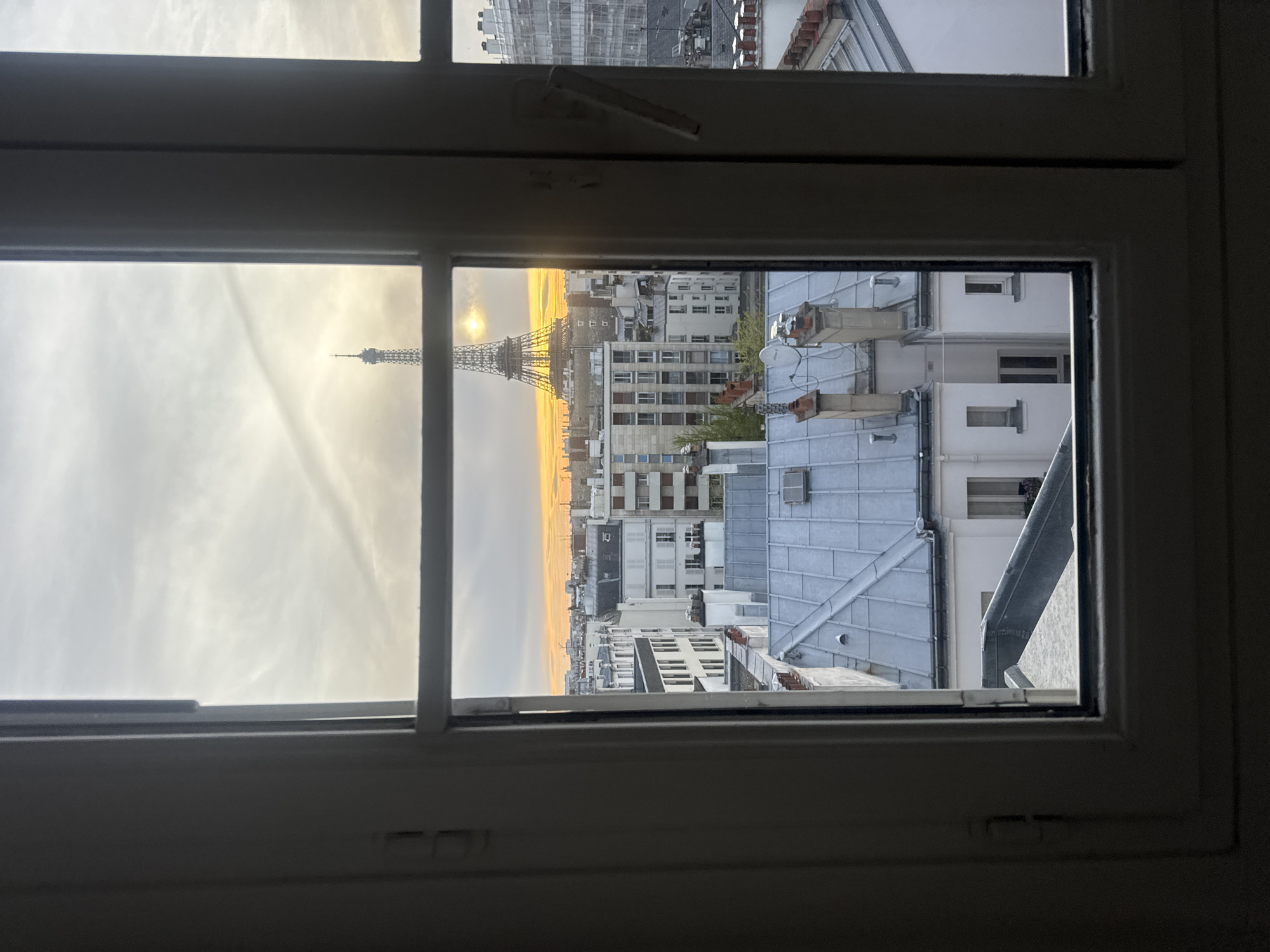 The image depicts a view of the Eiffel Tower through a window in Paris, France. The tower is visible in the distance, framed by the window panes, with the rising sun peeking from behind it. Other Parisian buildings with traditional architecture fill the space between the window and the Eiffel Tower. The window itself is white and partially open, suggesting the scene is viewed from inside an apartment. The sky has a sunset glow. The overall scene is still and peaceful.