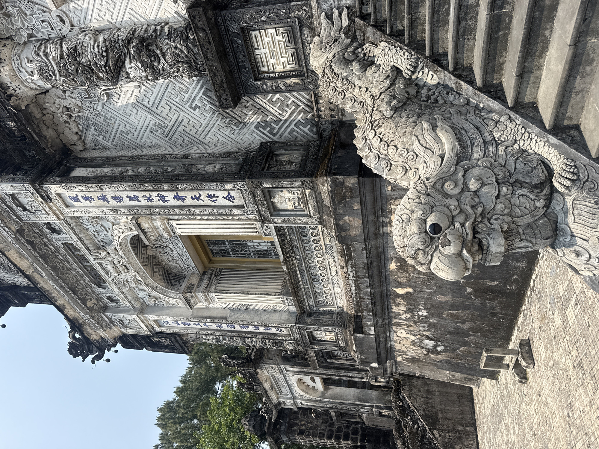 The image depicts a corner of a historic building, possibly a temple or palace complex, likely located in the city of Thủy Xuân Ward, Vietnam, judging by the architectural style and cultural details. The structure is characterized by intricate stone carvings, particularly a dragon-like figure that serves as a decorative element along a staircase. The building exhibits a blend of textures and patterns, with a prominent maze-like design on the ceiling and detailed inscriptions in a traditional script on the walls. A window with a lattice design can be observed along the façade. The overall ambiance suggests a place of historical and cultural significance, with the stone surfaces showing signs of age and weathering. Vegetation can be seen in the lower part of the image, hinting at an outdoor environment.