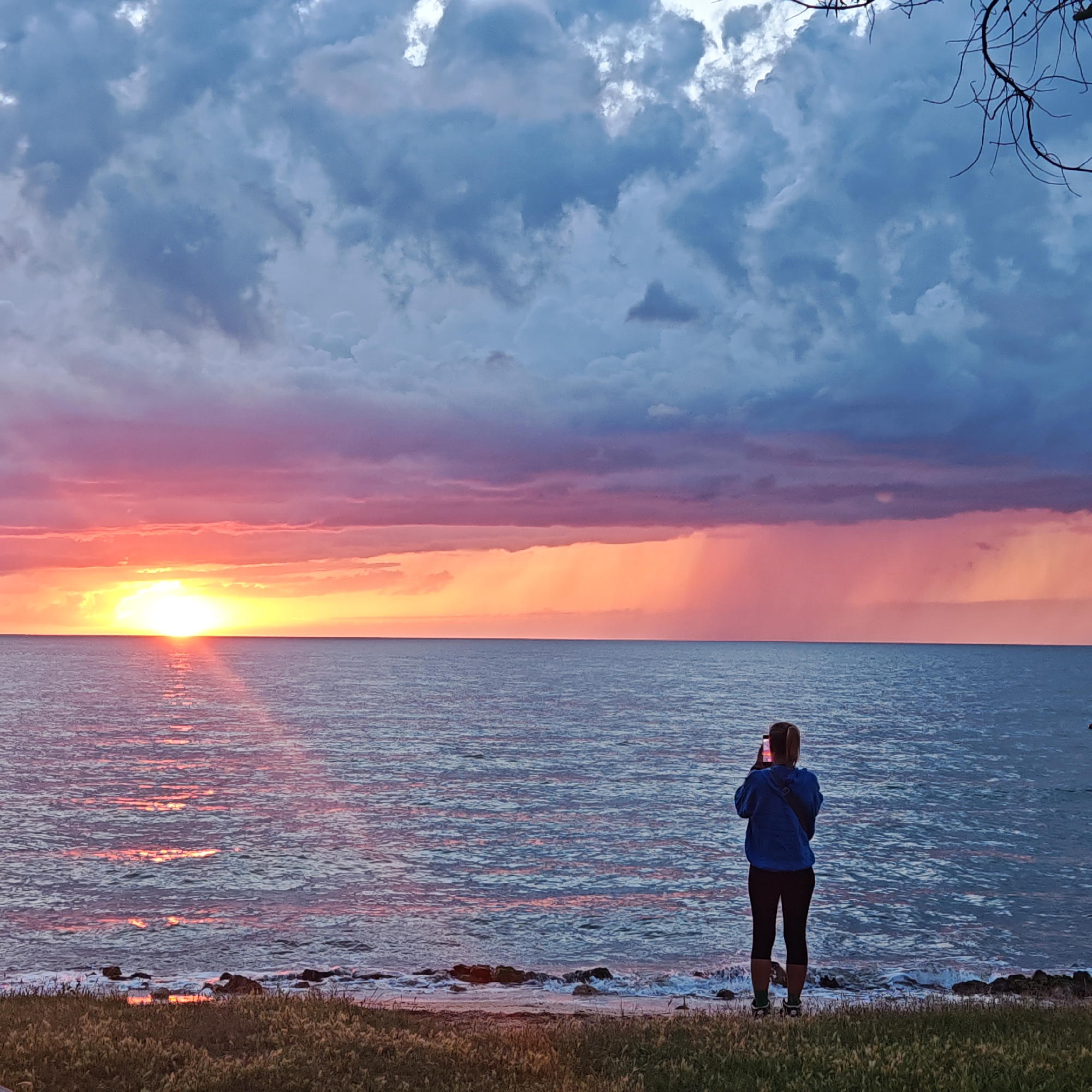 An image depicts a woman standing on a grassy shore, facing a body of water during a sunset. The sky is filled with dramatic clouds, ranging from dark blues to vibrant pinks and oranges, with rays of light breaking through. The sun is low on the horizon, casting a bright reflection on the water's surface. The woman is holding a phone up to take a picture of the sunset. The foreground shows dry grass and some rocks along the water's edge. The image location is specified as Chelyadinivske rural hromada, Ukraine.