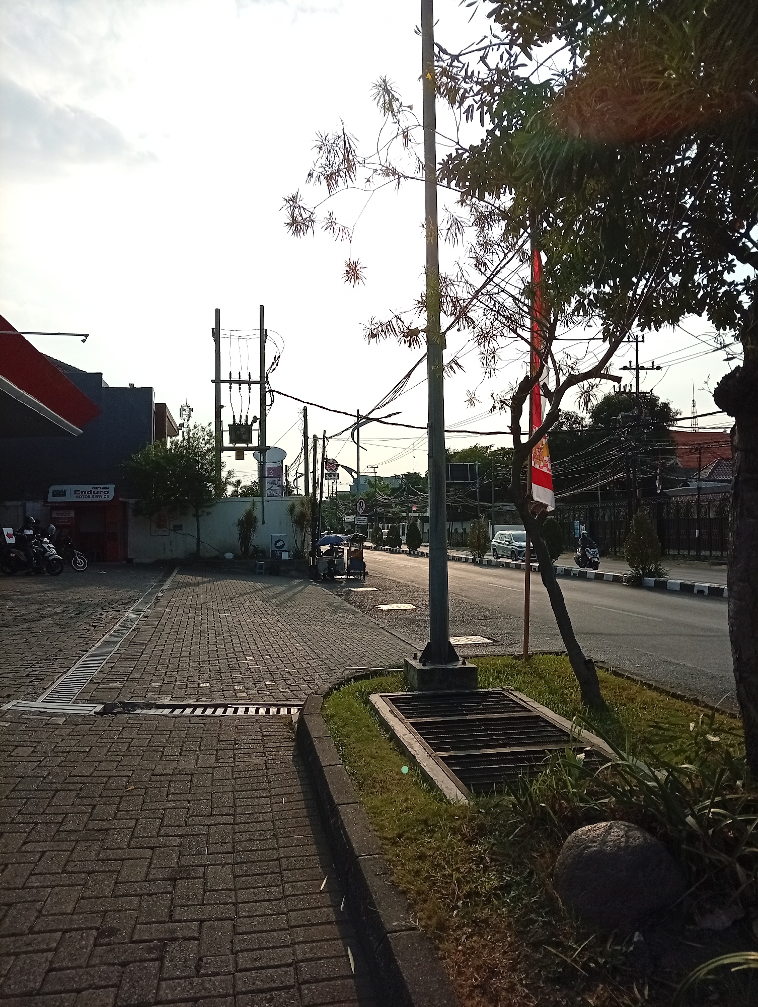This is a low-angle, full-shot photograph of a street scene in Surabaya, Indonesia, taken at dusk. The foreground is dominated by a cobblestone walkway with a metal grate drain. To the left, a red-roofed structure, possibly a gas station or shop, is visible with motorcycles parked outside. In the middle ground, a utility pole stands tall, adorned with a Indonesian flag. Across the street, a paved road runs with traffic, including cars and motorcycles. The background features buildings, trees, and an overcast sky, with the setting sun casting long shadows. The overall atmosphere is urban and slightly hazy.