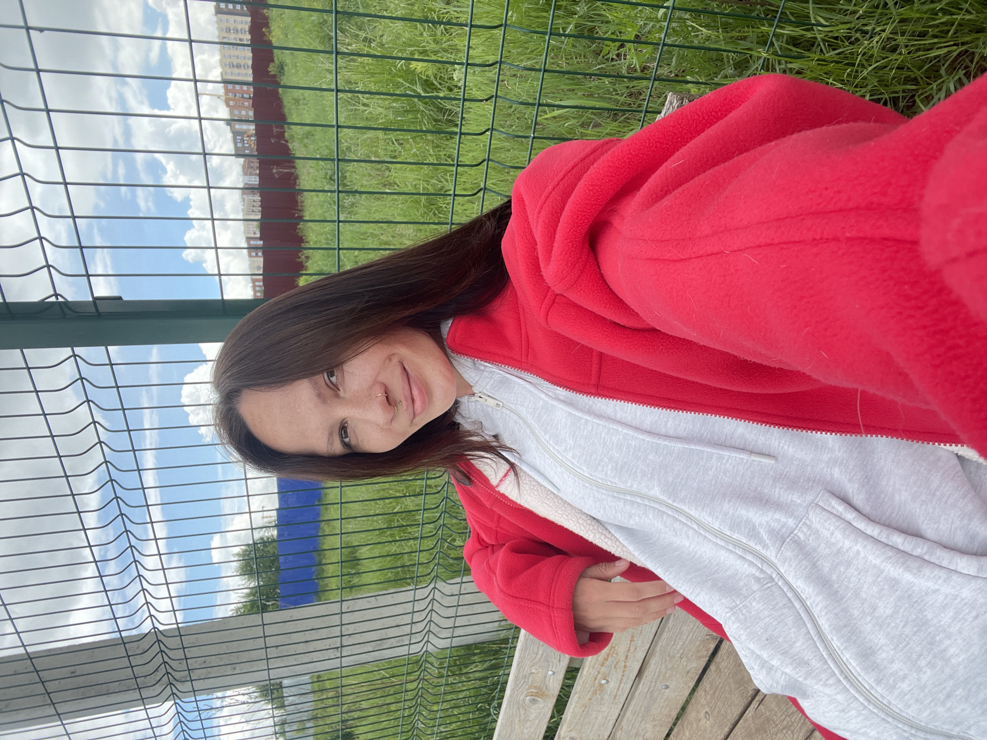 A young woman is depicted sitting on a wooden bench in an outdoor, daytime setting, likely in Izhevsk, Russia. She is looking towards the camera with a gentle smile. Her long, dark brown hair cascades over her shoulders, and she has a subtle nose hoop piercing. She is dressed in a vibrant red fleece jacket, worn over a light grey hooded sweatshirt. The sweatshirt has a visible white, possibly fleece, lining around the collar and front opening.
Behind her, a dark green metal fence with a grid pattern separates the immediate area from a expanse of green grass. Further in the background, under a bright blue sky with scattered white clouds, are several multi-story apartment buildings. The scene suggests a relaxed moment in a public space, such as a park or residential courtyard, on a clear, sunny day. No visible text is present in the image.