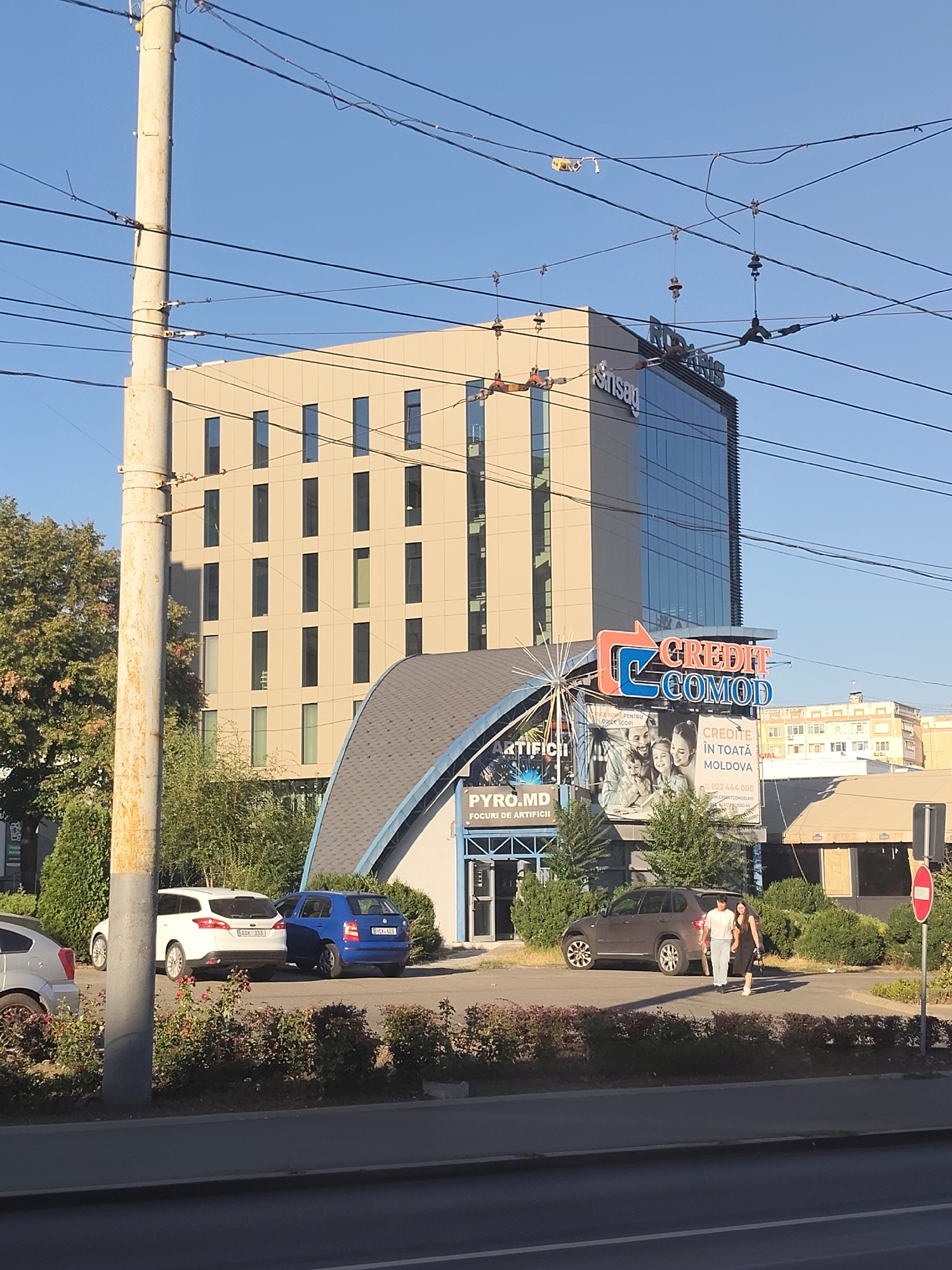 The image depicts a street scene in Chișinău, Moldova, on a clear day with blue sky. In the background, a modern, multi-story building with a beige facade and glass windows is prominent. Above the glass facade, the text "Sinsag" is visible. In front of this building, a smaller, curved-roof structure is situated. A large advertisement for "CREDIT. COMOD" is displayed, featuring images of people and text indicating services are available throughout Moldova. Below this, another sign reads "PYRO.MD" and "FOCURI DE ARTIFICII," suggesting a fireworks business.
Several vehicles are parked in front of the structures, including a white SUV and a blue car. Further to the right, a dark-colored SUV is parked, and two individuals, a man and a woman, are walking across the pavement. In the foreground, a section of a street is visible, with a concrete barrier separating it from a landscaped area with bushes. A tall, weathered utility pole with numerous wires runs vertically through the left side of the frame. The overall environment suggests an urban commercial area.