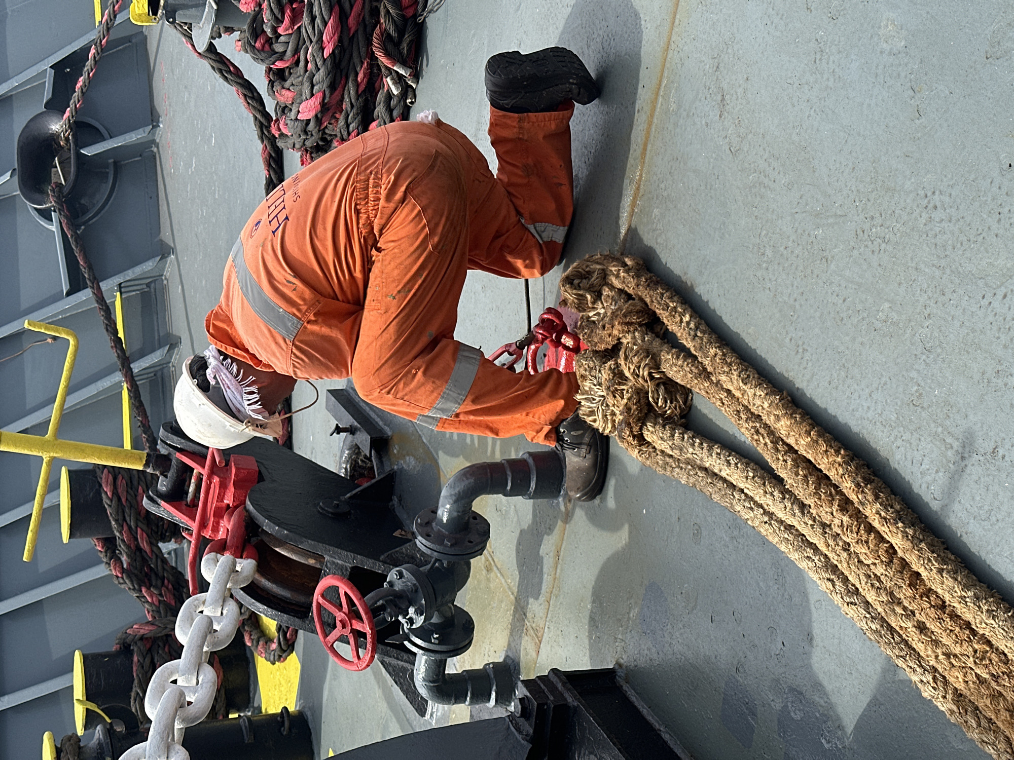 The image captures an individual in orange coveralls and a white hard hat, kneeling on the grey deck of a ship, specified as being in the city of Uribia, Colombia. The person is observed from the rear-right, bending over and attending to a piece of red and black machinery, which appears to be part of a ship's mooring or anchor handling system. This machinery features a large black drum or wheel, a red lever or brake mechanism, and a white anchor chain partially wrapped around it.
To the left of the kneeling person, a grey pipe system with a red valve wheel and bolted flanges extends across the deck. In the foreground, a thick, light brown, braided rope, heavily textured and appearing aged, lies horizontally across the deck, extending out of frame to the left.
In the background, behind the individual, several black mooring bollards with yellow caps are visible, around which thick black ropes with red intertwined strands are coiled. One of these ropes feeds into a black fairlead (mooring pipe) integrated into the ship's superstructure. The person's orange coveralls feature reflective strips on the legs and back, and a "HULL SHIPPING" logo is visible on the upper back. A white cloth or band is tied around the person's head beneath the hard hat. The deck surface appears textured and slightly damp in areas, with visible wear and tear. The overall environment is industrial, outdoors, and under daylight conditions.