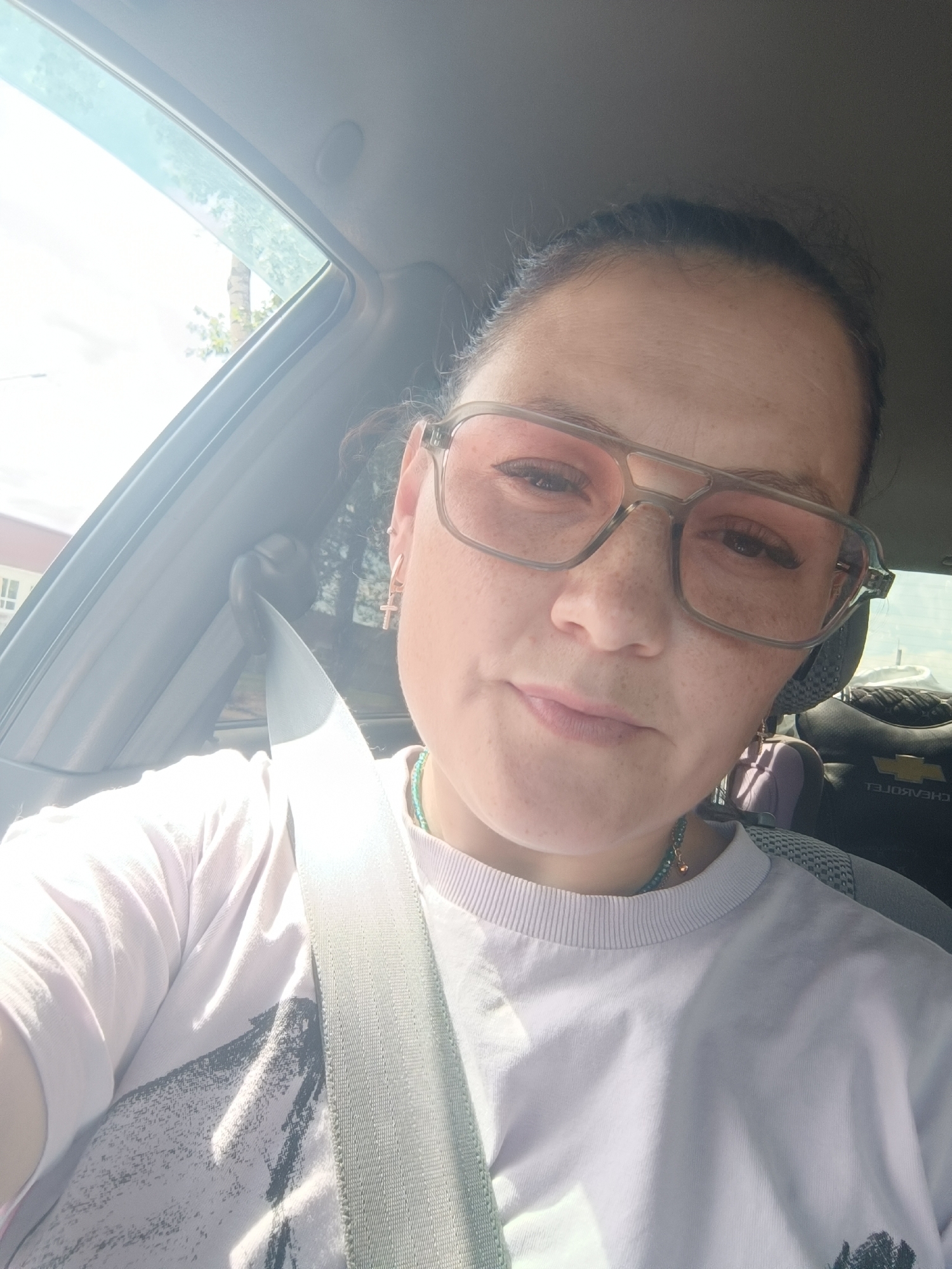 A woman is seen from the chest up, taking a selfie in a car. She is wearing pink-tinted aviator glasses and a light-colored t-shirt with a graphic print. A seatbelt is visible across her chest. The background shows the interior of a car, including a headrest with a Chevrolet logo. Sunlight streams in from the left, illuminating the scene. The image appears to be taken during the daytime, outdoors, possibly in a residential area with some trees visible outside the car window. There are no visible indications of the specific location within Chaikovsky, Russia, other than it being a general urban or suburban environment.