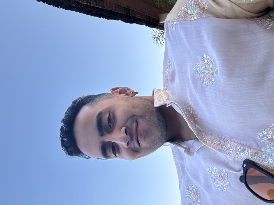 A low-angle close-up photograph depicts an adult male in the city of Malasar, India, positioned against a clear blue sky. The man, of apparent South Asian descent, has short dark hair and light facial stubble. He is smiling directly at the camera. He wears a light-colored, possibly pale pink or off-white, traditional Indian garment, likely a kurta or sherwani, featuring a band collar and intricate embroidery with pastel-colored threads and reflective accents across the chest and collar. A small, gold-colored hoop earring is visible in his right earlobe. A pair of black-framed sunglasses with amber-tinted lenses are hooked onto the front of his garment below the chest. In the upper right background, a portion of a thatched roof structure and sparse green foliage are visible.