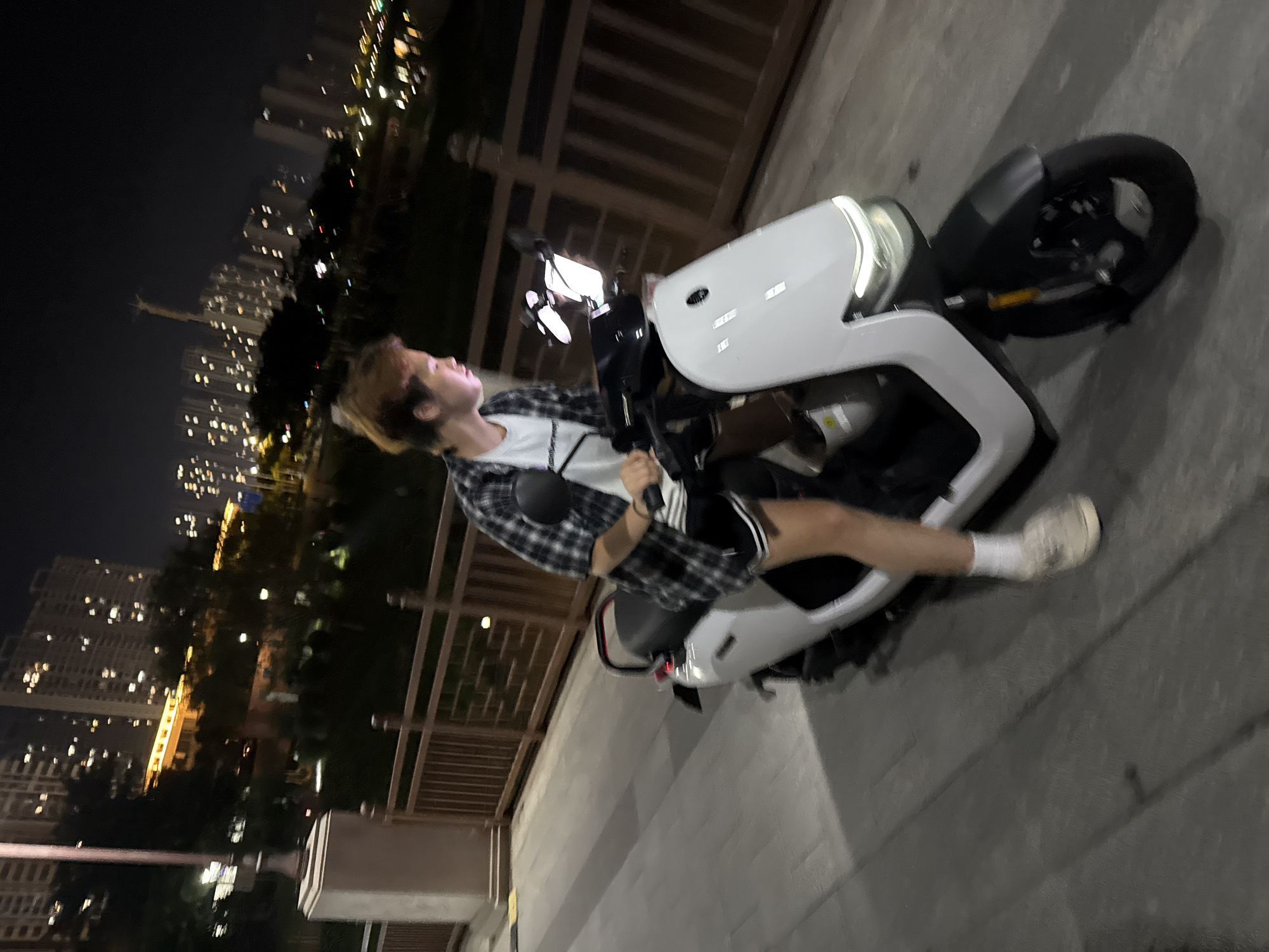 A person with blondish-brown hair, wearing a white t-shirt and a plaid shirt over it, along with black shorts and white socks with light-colored shoes, is riding a white electric scooter at night. The scooter has a white body and a black front fender and wheel. The person's face is partially illuminated by the scooter's headlight. They are on a paved walkway with a wooden railing to their right, which is adjacent to a body of water. In the background, there are tall buildings with many illuminated windows, suggesting an urban setting. The lighting is dim, indicative of nighttime. The overall scene appears to be a waterfront area in Huizhou, China, likely a park or promenade, given the presence of the walkway, railing, and the cityscape in the distance.
