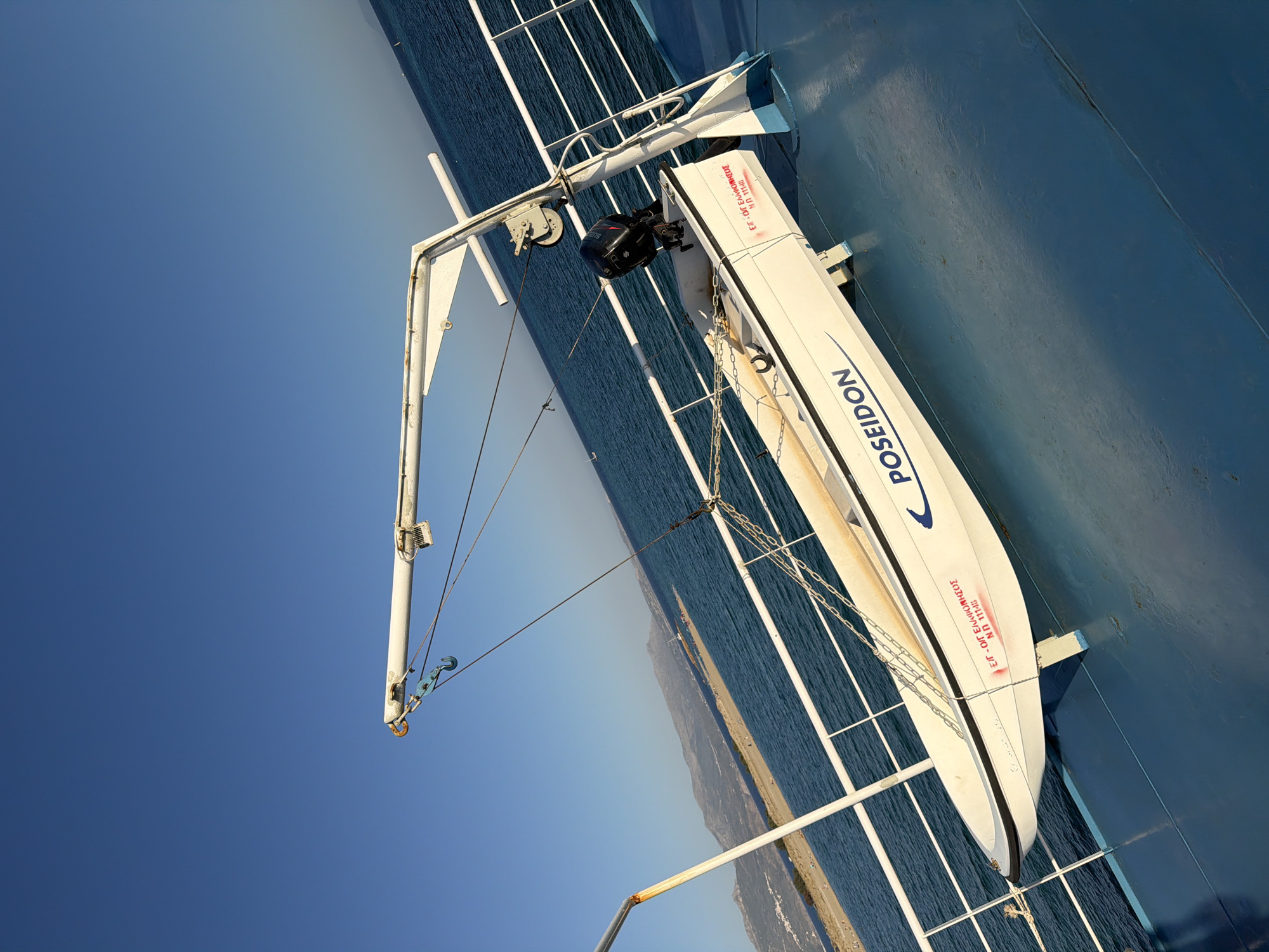 The image depicts a life raft, labeled "POSEIDON", secured by a crane on the deck of a ferry. The ferry's deck is painted blue, and a white railing runs along its edge. The sea is visible in the background, with a beach and mountains on the coast of Elafonisos, Greece. The sky is clear and blue. The scene appears to be a sunny day, suggesting a calm environment. There are no people visible in the image. The life raft has an outboard motor attached to its stern. Red lettering is present on the side of the life raft, but the text is partially obscured. The crane is a white metallic structure, with cables and pulleys visible.