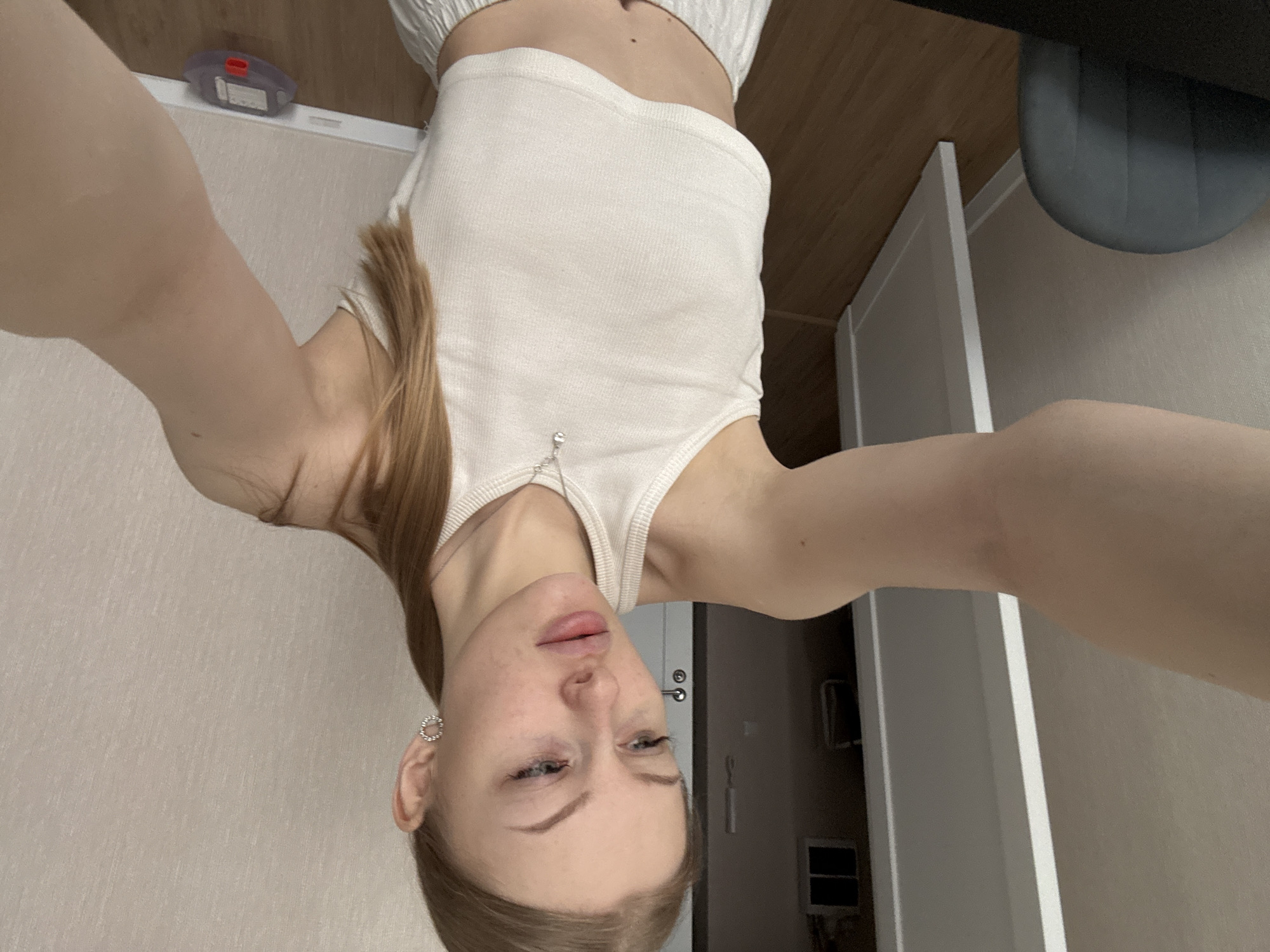 This media file features a selfie of a young woman taken in an indoor setting, presumably in Perm, Russia. The image, which is originally oriented upside down, captures the woman from a high angle, looking down towards the camera she holds with extended arms.
The subject is a young woman with light brown hair, which appears to be pulled back. She is wearing a simple white ribbed crop top. Her facial expression is neutral, with slightly parted lips and light-colored eyes. She wears small, circular, silver-toned earrings.
The background shows a minimalist room interior. On the left side of the frame, a light-colored wall meets a dark wooden floor. Resting on the floor near the baseboard is a purple and red inhaler, with some red and white text partially visible on its surface. To the right, a white door frame is prominent, and a section of a dark grey object, possibly a chair or stool, is visible in the upper right corner. The lighting is soft and even, suggesting a calm indoor environment. The image conveys a casual, personal moment.