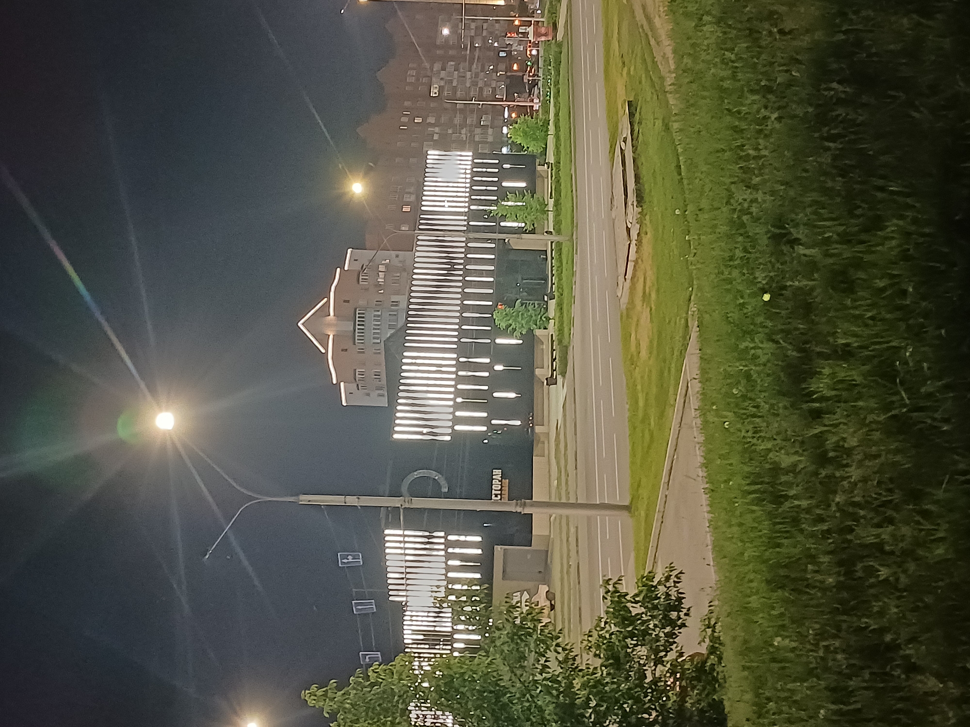 The image depicts a nighttime scene in an urban environment. In the foreground, a patch of lush green grass and a paved area are visible. A street runs horizontally across the middle ground, with a traffic light visible on the right side. Beyond the street is a sidewalk and a modern building with a dark facade and numerous vertical white lights. The building features a large circular logo on its facade and the word "СТОРАН" in orange letters. A tree with green foliage is situated on the left side of the building. In the background, several apartment buildings are illuminated. A streetlamp with a bright light shines in the upper left quadrant of the image. The sky is dark and clear.