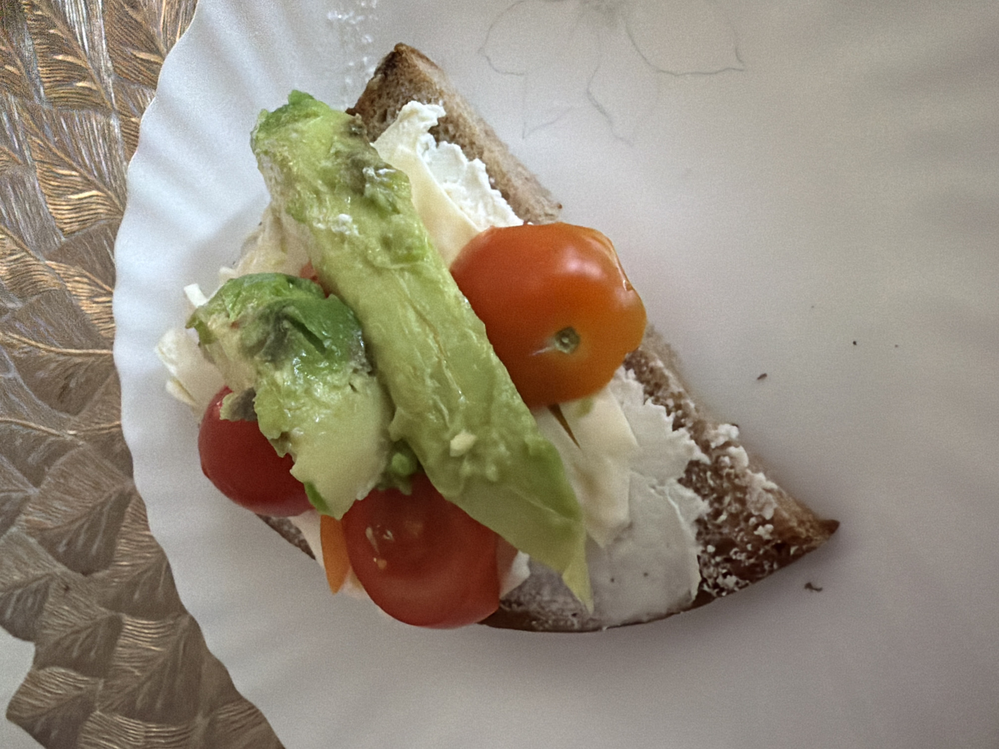A sandwich rests on a white paper plate. The sandwich is constructed on a triangle of toasted brown bread spread with a white creamy substance, possibly cream cheese or mayonnaise. Toppings include slices of avocado, small red and orange cherry tomatoes, and shredded lettuce. The plate rests on a surface with a leaf-patterned texture. The sandwich is centrally located in the frame. The specific location in Budva, Montenegro is not evident in the image.