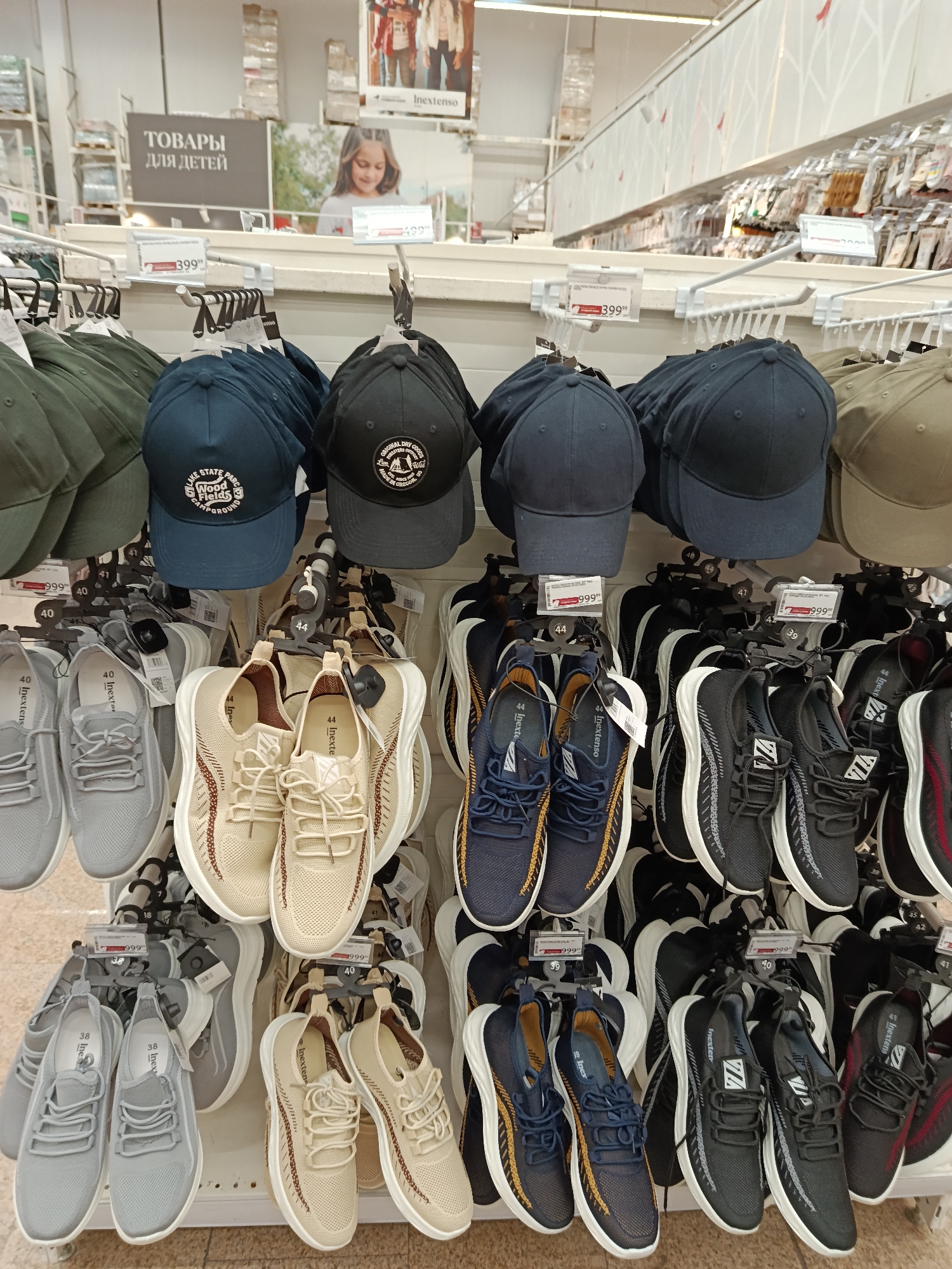 The image shows a shelf in a store, possibly in Moscow, Russia, displaying clothing items. On the top shelf, there are several baseball caps in varying colors: green, blue, black, and khaki. Some of the caps have logos or writing on them, such as "LAKE STATE WOOD Fields CAMPGROUND" and "ORIGINAL DRY GOODS." Below the caps, there are rows of sneakers hanging by their laces. The sneakers come in shades of gray, beige, navy blue, and black, with white midsoles. Each pair of sneakers has a tag indicating its size. There are price tags displayed near the merchandise indicating the cost of items, which are mostly 399 and 999. In the background, there is a sign that reads "ТОВАРЫ ДЛЯ ДЕТЕЙ" (Products for Children) and an advertisement featuring a girl. The overall impression is that of a clothing section in a retail store.