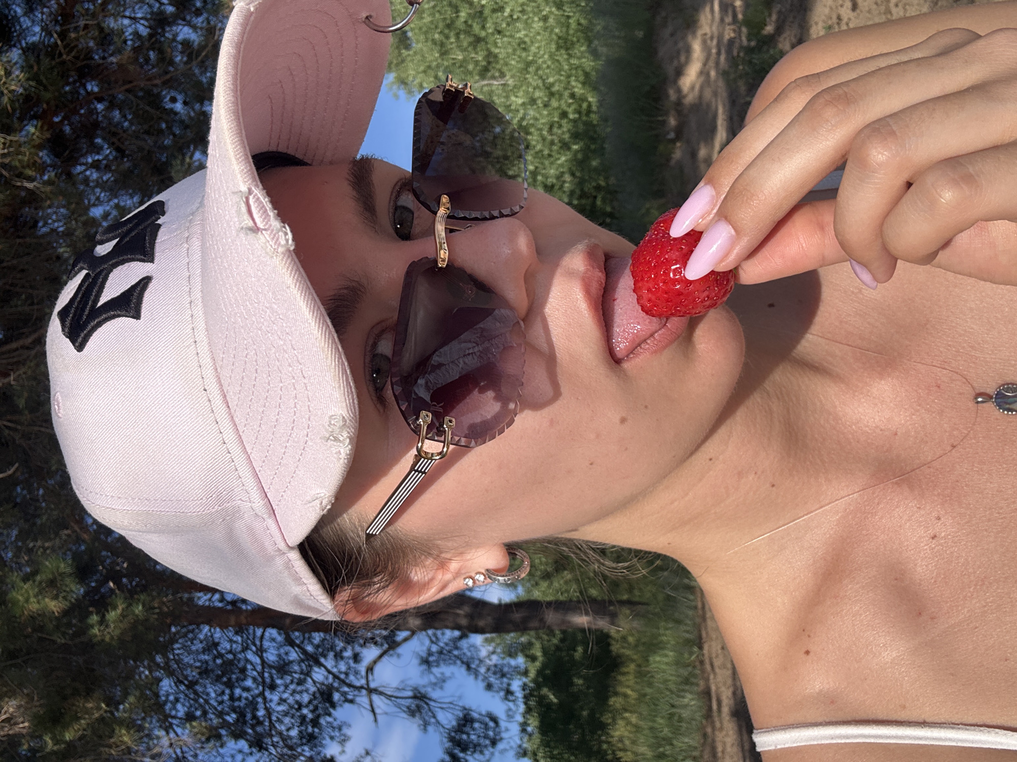 A young woman wearing a pink baseball cap and sunglasses is eating a strawberry in a park on a sunny day. She is holding the strawberry with her right hand and licking it with her tongue. The woman has light skin and long dark hair. The background is blurred with trees and grass.