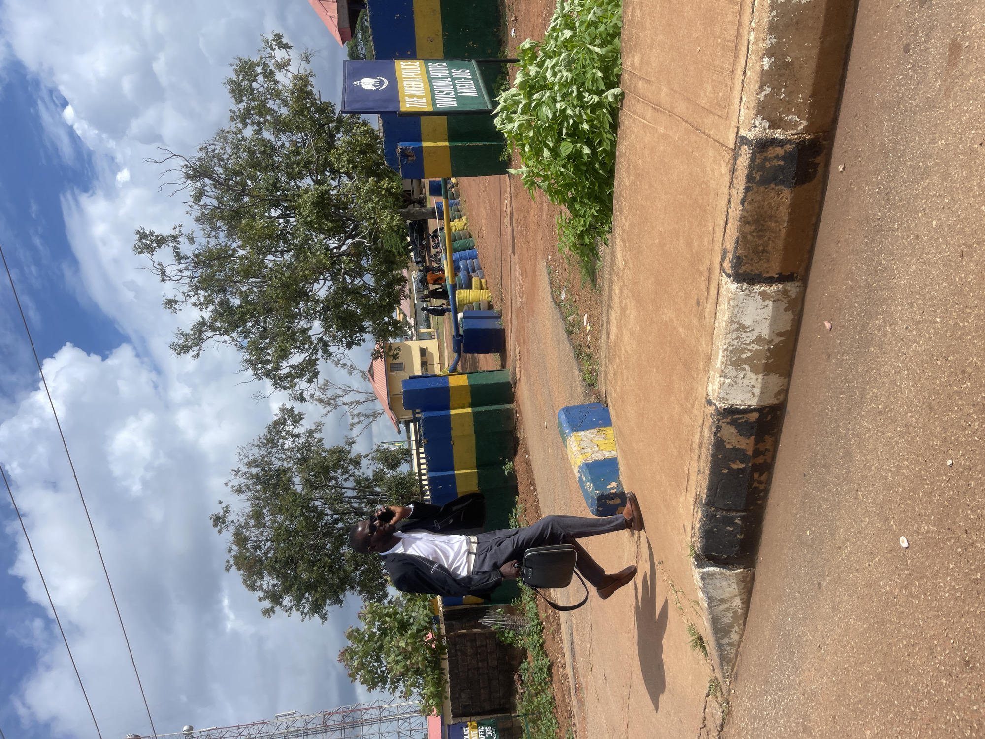 A Black male in a suit, white shirt, and sunglasses is walking on a dirt road, talking on a cell phone. He is carrying a black briefcase in his left hand. The road is bordered by a concrete curb painted with black and white stripes. Behind him is a building with yellow and blue painted walls, a sign that reads "THE NIGERIA POLICE DIVISIONAL HQTS ANGIO JOS," and a large tree. The sky is cloudy with patches of blue. The scene appears to be in a semi-urban or rural area in Nigeria.
