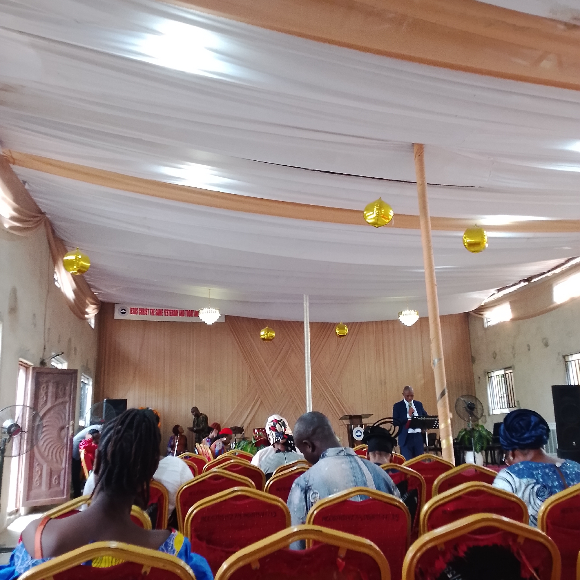 The image depicts an indoor gathering, likely a church service, within a venue in Ado, Nigeria. The space is decorated with draped white and gold fabric lining the ceiling and walls, creating a festive atmosphere. Several gold, spherical balloons are suspended from the ceiling, adding to the celebratory feel.
In the foreground, the backs of several congregants are visible, seated in rows of red chairs with gold trim. Their attire suggests a formal or semi-formal occasion, with some wearing headscarves. The focus of the gathering appears to be a speaker at a lectern towards the back of the room, dressed in a suit. A banner above the speaker reads, "JESUS CHRIST THE SAME YESTERDAY AND TODAY AND FOREVER." Other elements visible include a chandelier-like light fixture, fans, and windows with blinds. The overall lighting suggests an indoor setting, possibly with natural light filtering in from the windows. The wooden paneling on the walls further contributes to the established ambiance.