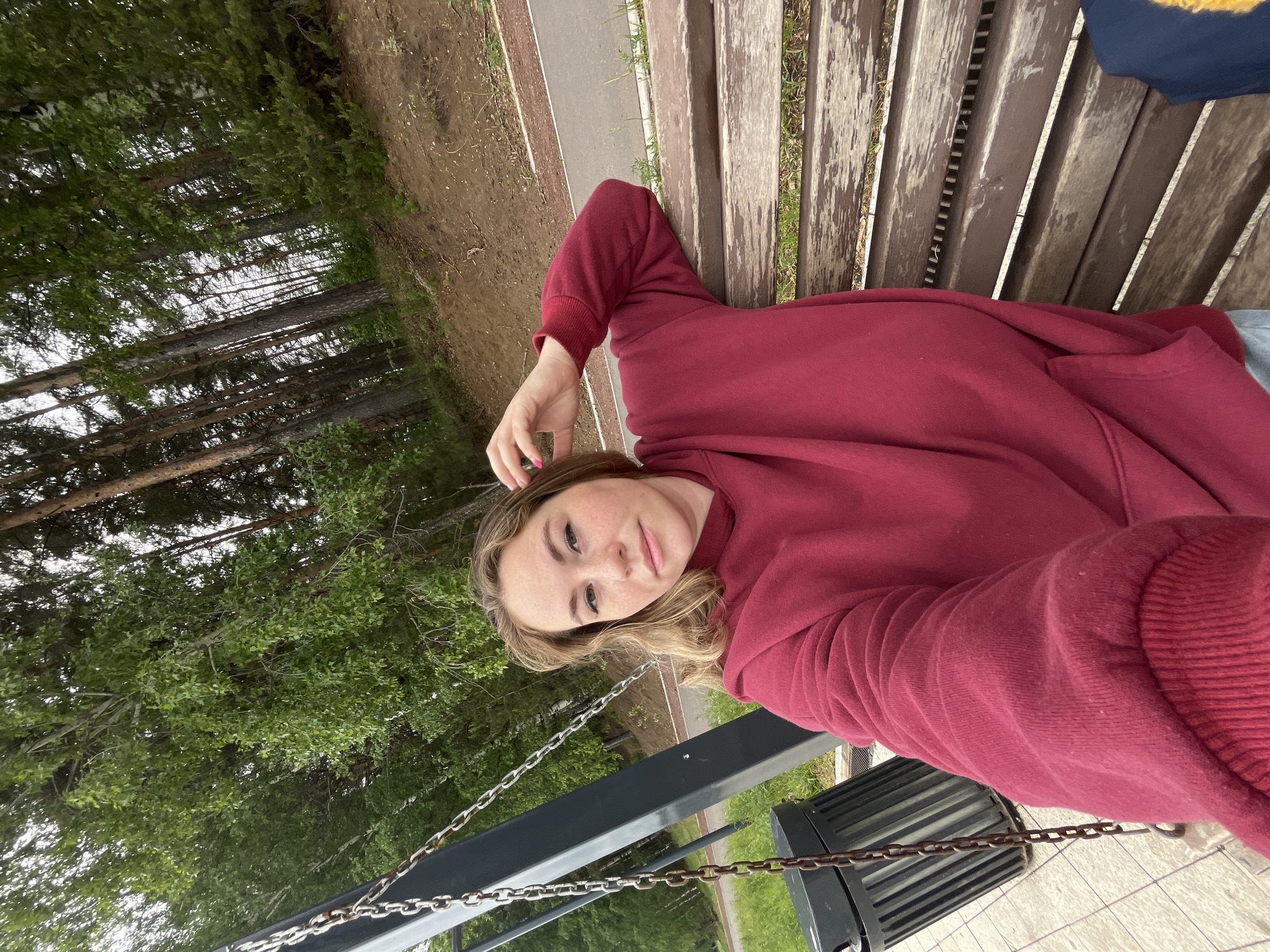 A young woman lies on a wooden park bench, wearing a red sweatshirt. She looks directly at the camera, her right hand near her head. Trees are visible in the background. The bench is situated near a path in the city of Chaikovsky, Russia. A black trash can is attached to the bench by metal chains.