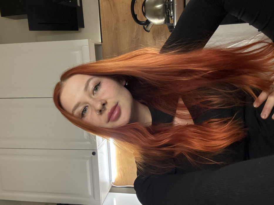 A light-skinned person with long, red-orange hair is positioned at a slight angle against a backdrop of kitchen features. Notable details include visible freckles, a nose ring, and full lips. The person is wearing a black top with long sleeves. In the background, there are white cabinets, a black box on the wall, a wooden countertop, and a metal kettle. The image appears to be taken indoors with ample lighting.