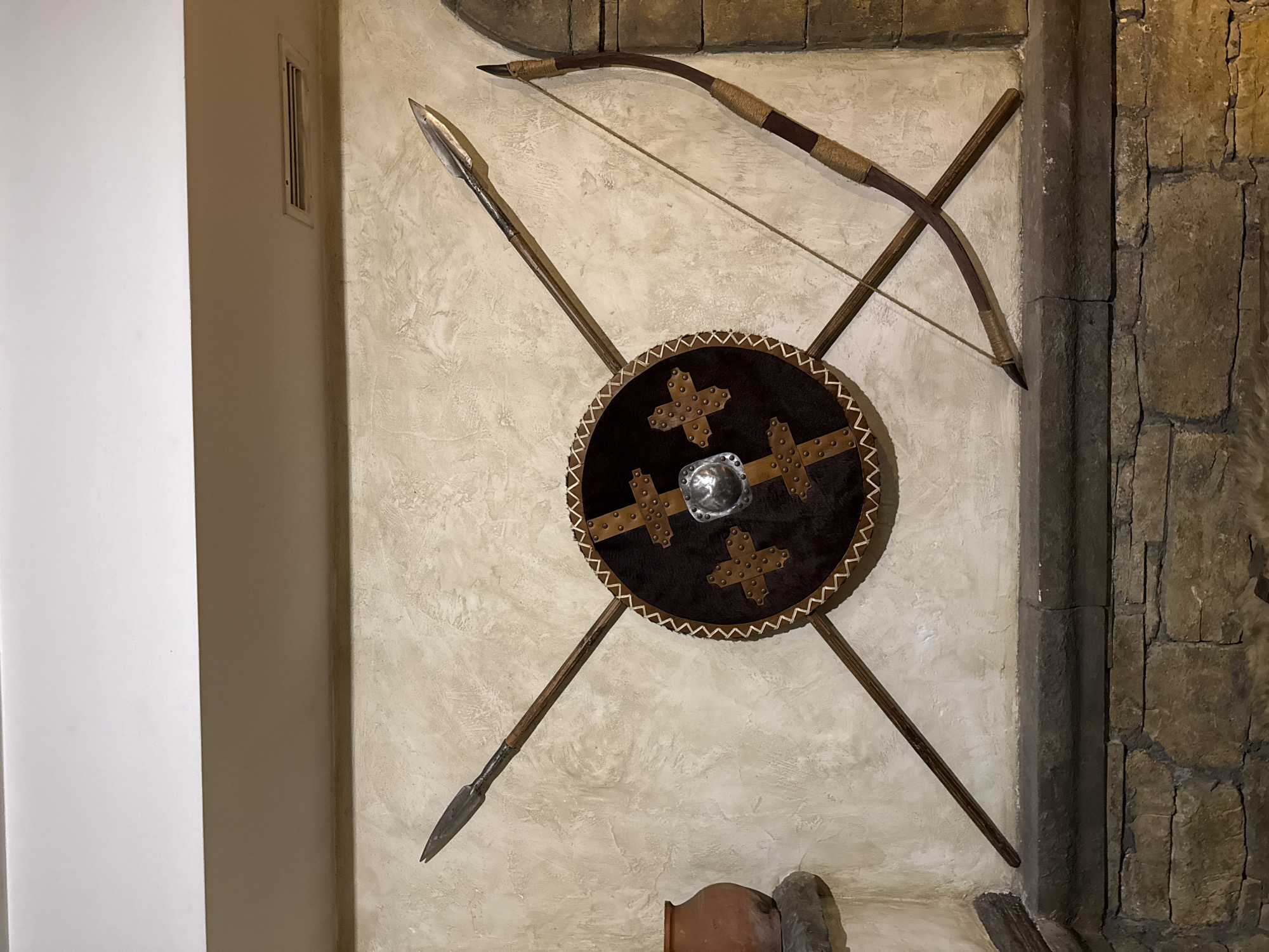 A still shot shows a wall arrangement of medieval weaponry. A circular dark brown shield with leather trim and studded crosses is centered. A spear and bow and arrow are diagonally crossed behind the shield. The wall has a textured, off-white finish, transitioning into a stone-like corner. The location is presumably Veliko Tarnovo, Bulgaria, known for its medieval history.