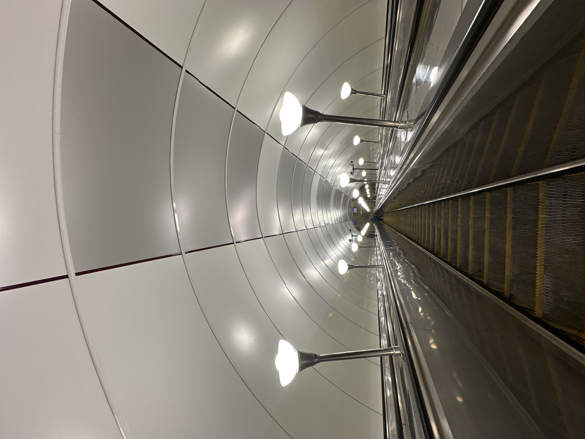 An upward-facing, wide-angle view of a long escalator in the Saint Petersburg Metro, Russia. The escalator has a textured yellow and black tread and polished metal sides. There are multiple modern, white, tulip-shaped lamps lining the right side of the escalator, which are illuminated. The ceiling is a series of white, curved panels with thin, dark red lines accentuating the curves. The perspective draws the viewer's eye towards a distant point where a few individuals, dressed in casual clothing, are visible at the top of the escalator. The scene conveys a sense of depth and the expansive nature of the metro station.
