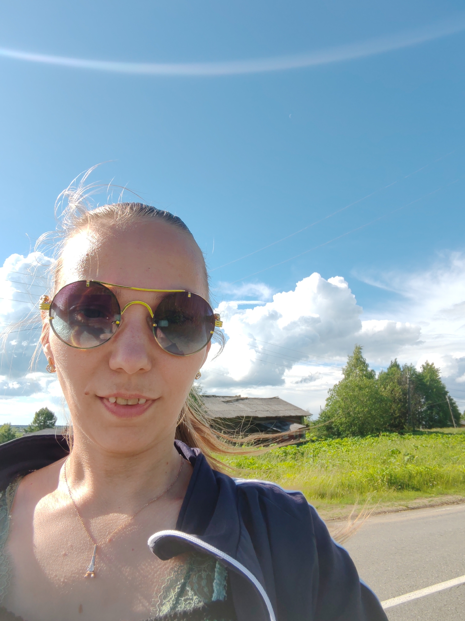 This is a close-up, outdoor selfie of a person wearing large, round, mirrored sunglasses with gold frames. The subject has blonde hair and is smiling slightly, showing some teeth. They are wearing a dark blue jacket over a green lace garment and a delicate necklace with a pendant resembling a rocket. The background suggests a rural setting in Russia, with a bright blue sky dotted with fluffy white clouds. A faint rainbow arc is visible in the upper left of the sky, indicating sunshine and possibly recent rain. In the mid-ground, there's a field of green grass and vegetation, a rustic wooden building with a corrugated roof, and several trees. A paved road is visible in the lower right corner. The time of day appears to be daytime, likely afternoon, given the bright sunlight and the clear blue sky. The wind is blowing, evident from the way the subject's hair is swept back. There is no visible text in the image.