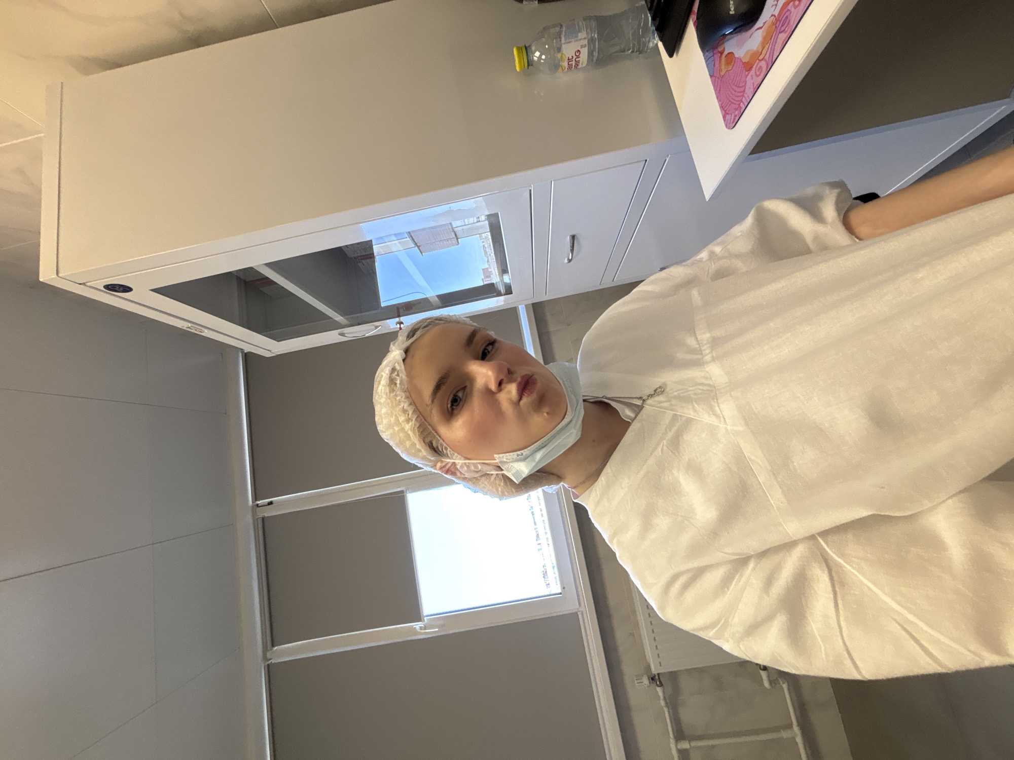 The image shows a woman in a white bouffant cap and surgical mask, wearing a white garment. The setting appears to be an indoor space, potentially a clinic or laboratory, with bright sunlight streaming through a window. A desk is visible, with a bottle of water, a computer mouse, and a mouse pad on top. The location is possibly in Saint Petersburg, Russia, inferred from the architectural details visible through the window.