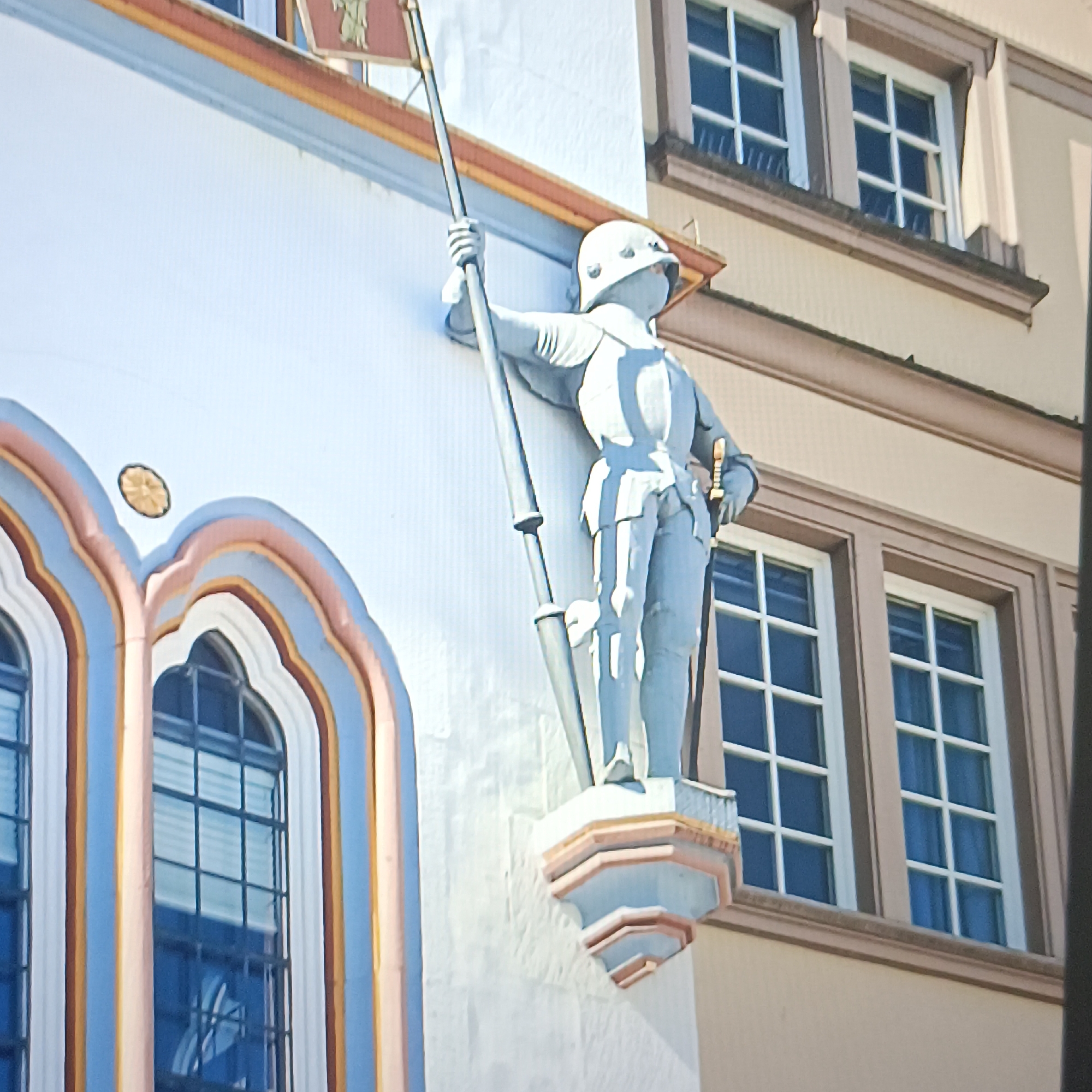 A statue of a knight in armor holding a spear and a sword stands against a light-colored building. The knight is depicted in a light blue color and is positioned to the left of the frame. Behind the knight, windows with white frames and dark blue panes are visible. The building has decorative architectural elements, including arched windows with colorful trim in shades of blue, pink, and yellow on the lower left side of the image. The upper part of the building features a red and gold banner with a symbol that is partially obscured. The overall scene appears to be an exterior view of a historical building, possibly in a European city.