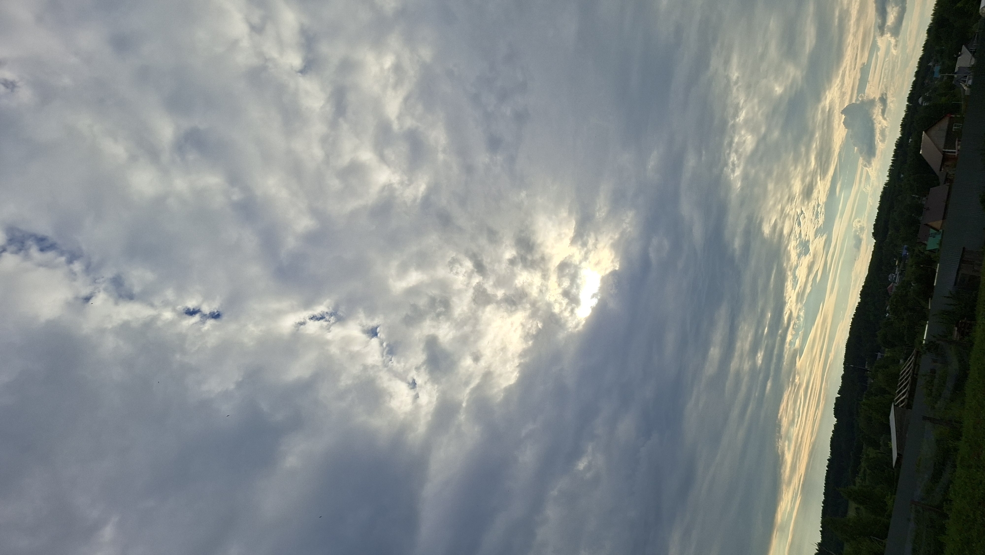 The image depicts a cloudy sky over a rural landscape in Большой Букор, Russia. The sky is filled with a mix of dark and light gray clouds, with patches of sunlight breaking through in the upper half of the frame. Below the sky, a dense line of green trees forms a treeline on a rolling hill. Scattered among the trees and on the slopes of the hill are several houses and rooftops, suggesting a small village or scattered settlement. In the foreground, a fence runs horizontally across the bottom of the image, with glimpses of grassy areas and some outbuildings. The overall scene appears to be an outdoor view captured during daylight hours, possibly late afternoon or early evening due to the lighting.