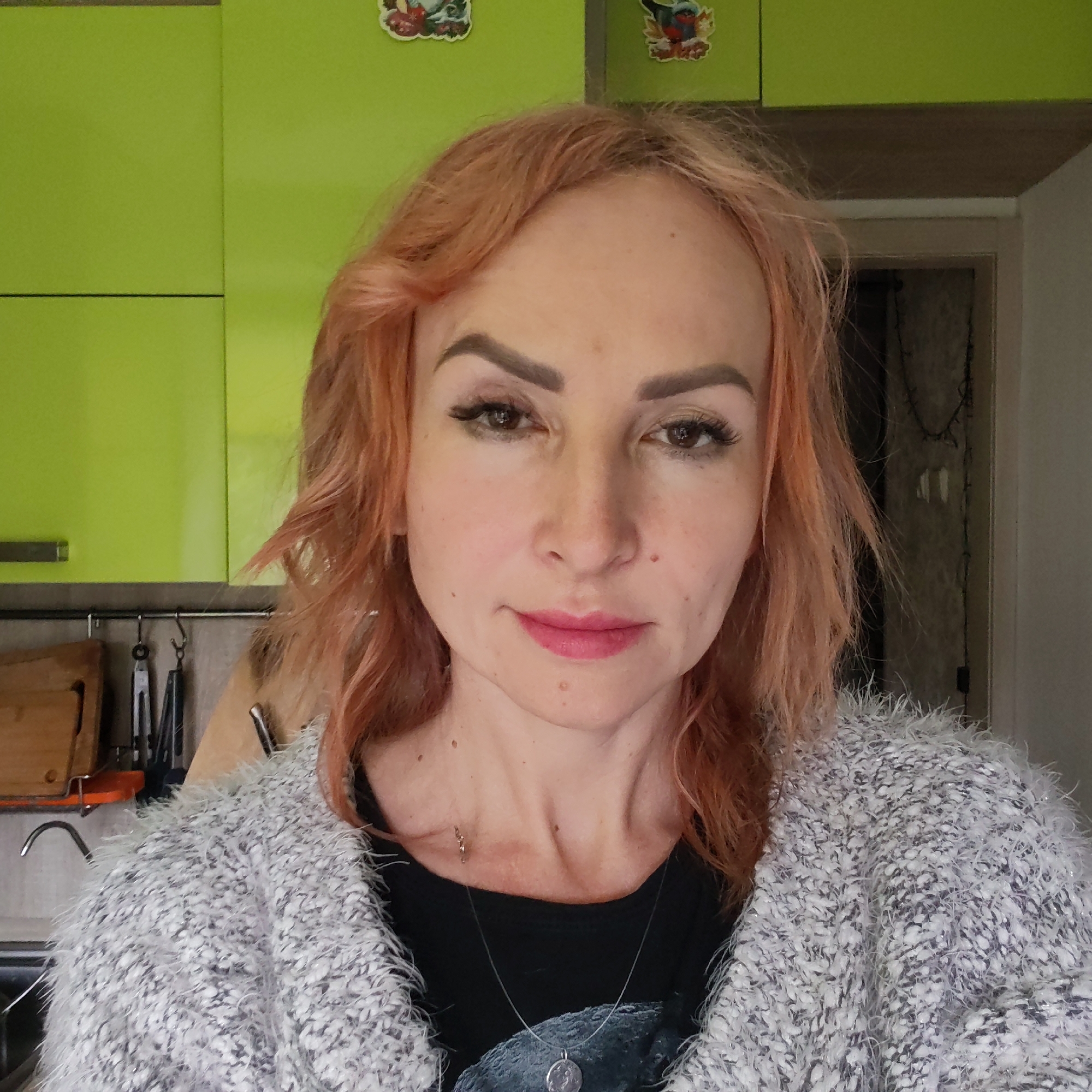 The image presents a head-and-shoulders portrait of an adult female, attributed to the city of Chaikovsky, Russia. The subject has reddish-orange, wavy, shoulder-length hair, fair skin with multiple moles or freckles, dark defined eyebrows, long eyelashes, and is wearing pink lipstick. She looks directly at the viewer with a neutral to slight smile expression. Her attire consists of a black round-neck top layered with a textured, fuzzy grey and white cardigan. A thin, silver-colored necklace with a circular pendant rests on her chest. The background on the left side features a kitchen area with bright lime green upper cabinets, a horizontal darker strip, and a metal rail holding several kitchen utensils. A wooden cutting board and a chrome faucet are partially visible on a countertop below. The background on the right side shows a darker wall area, possibly a doorway or hall, with some indistinct wire-like decorations and a glimpse into a darker room. Two small, colorful decorative magnets are attached to the upper green cabinet.